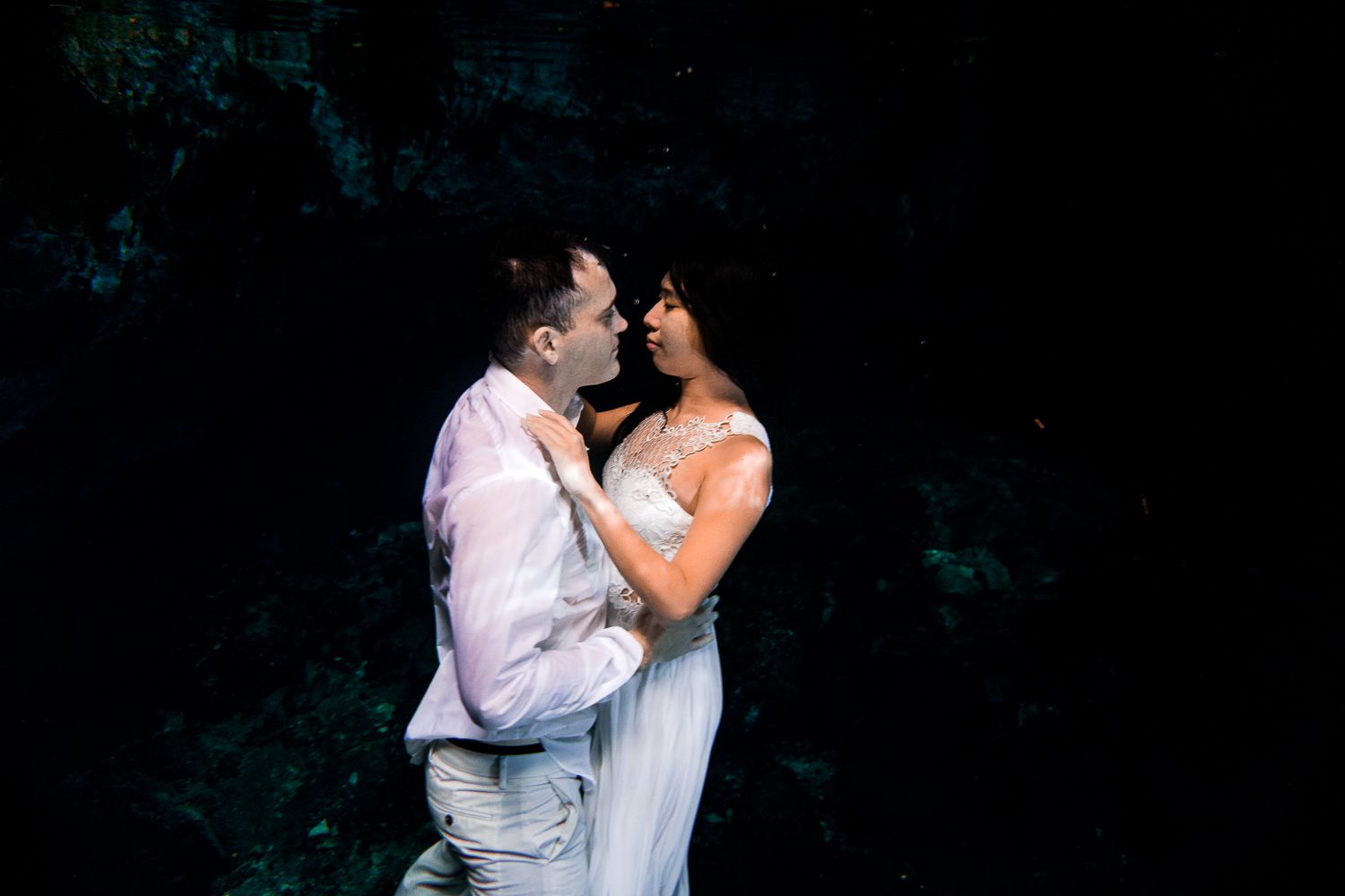 Couple in wedding attire embrace in dramatic dark cenote underwater setting.