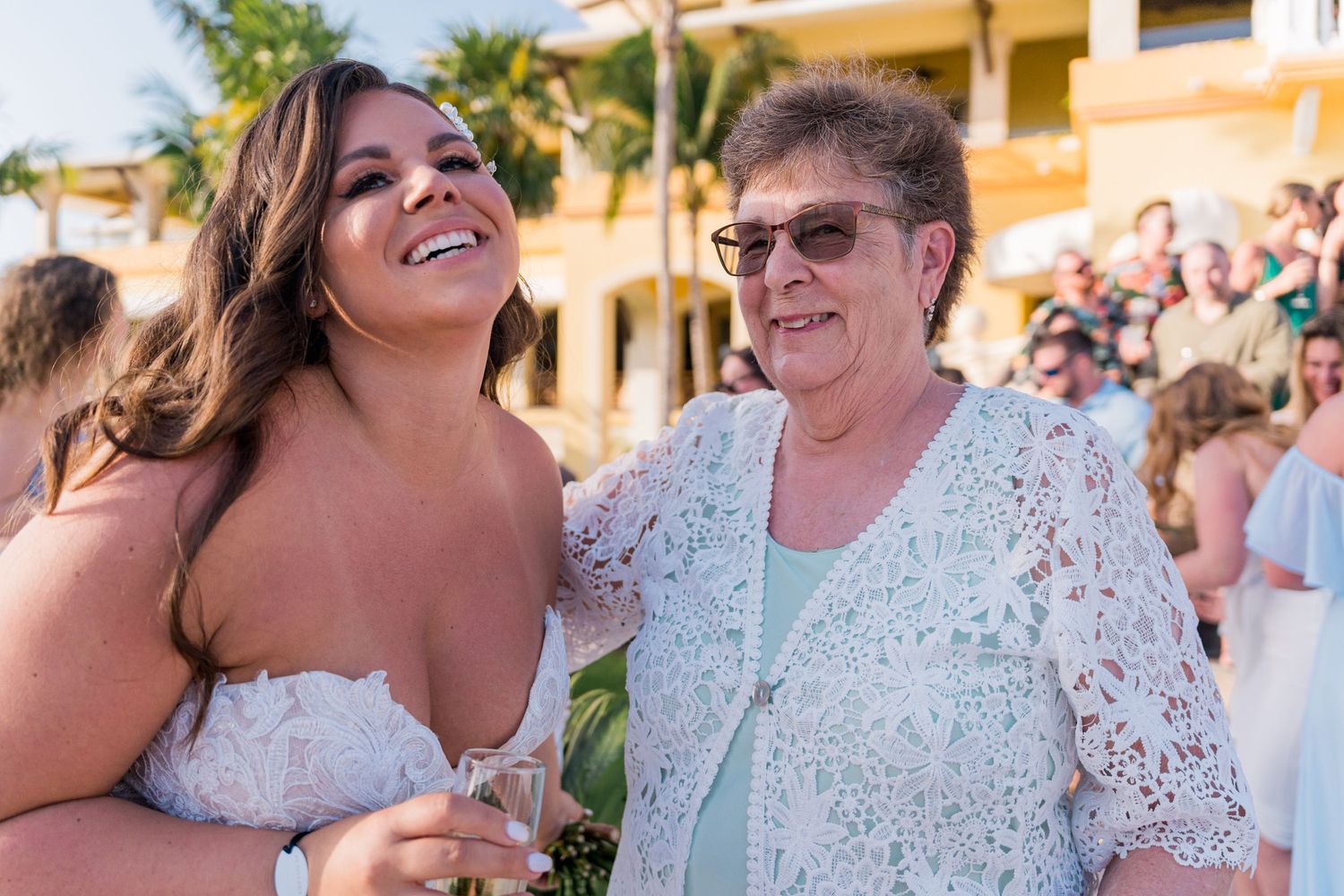 Bride laughing with an elderly woman in a white lace dress at a sunny outdoor wedding.