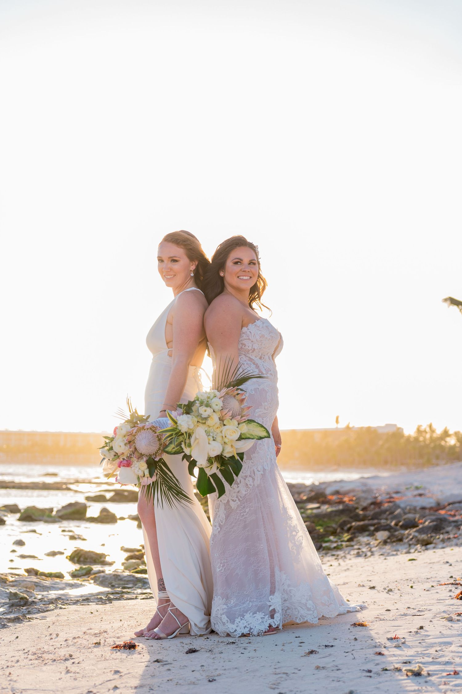 Two brides standing back-to-back on a beach holding floral bouquets in golden sunset light.