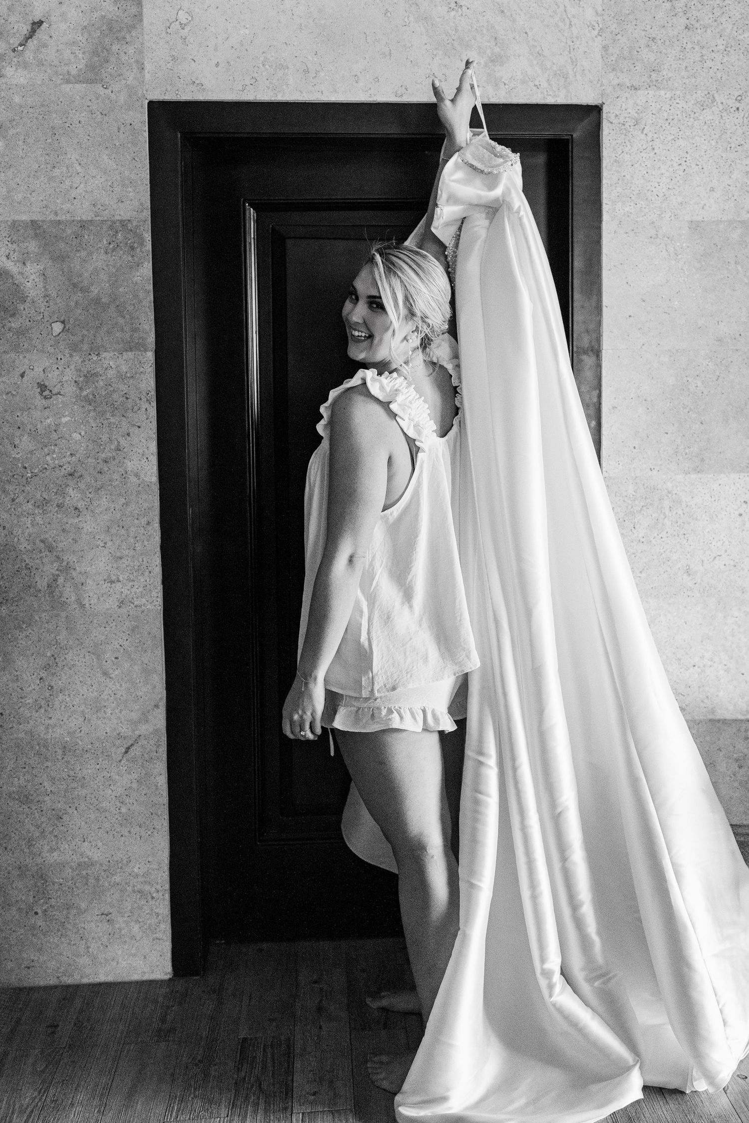 Black and white photo of a smiling bride holding her wedding gown in a doorway wearing a white slip.