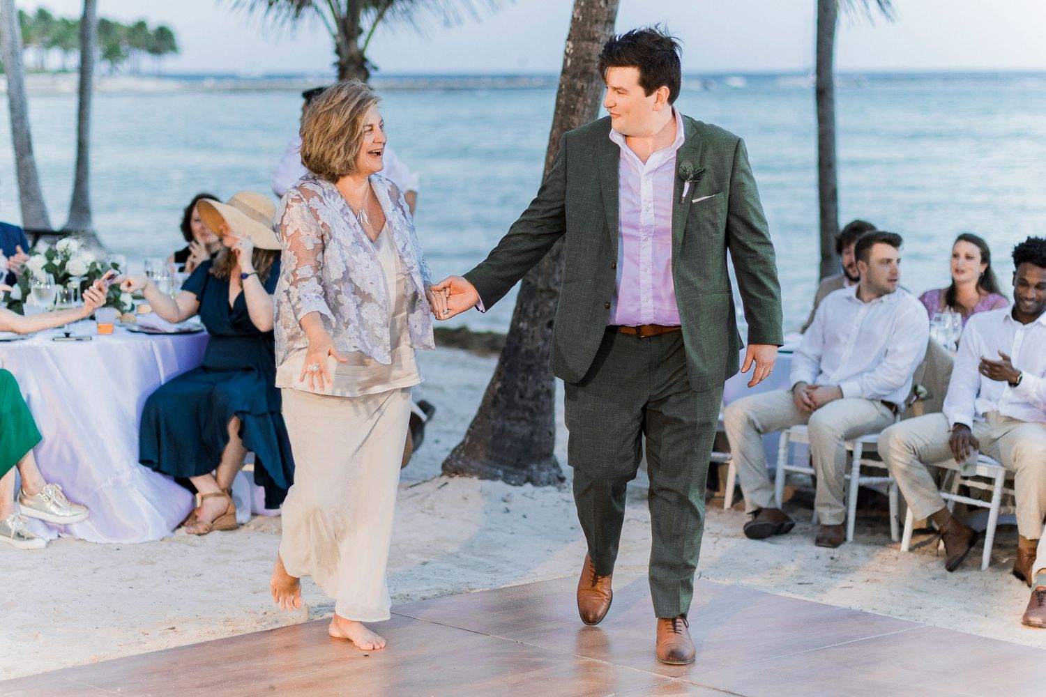 Couple holding hands dancing on beach wedding reception dance floor with guests seated under palm trees at sunset.