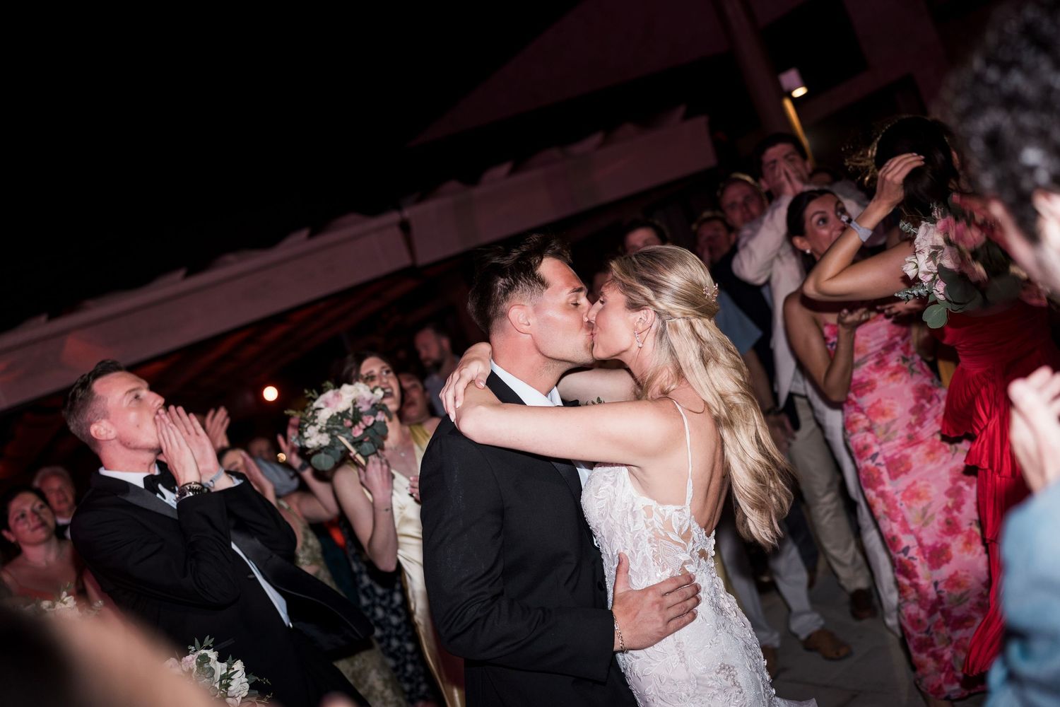 Bride and groom share a romantic kiss surrounded by cheering guests at an outdoor evening wedding reception.