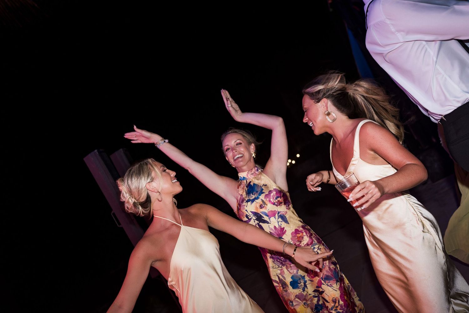 Three women in elegant dresses dancing and laughing together at an outdoor evening celebration or wedding reception.