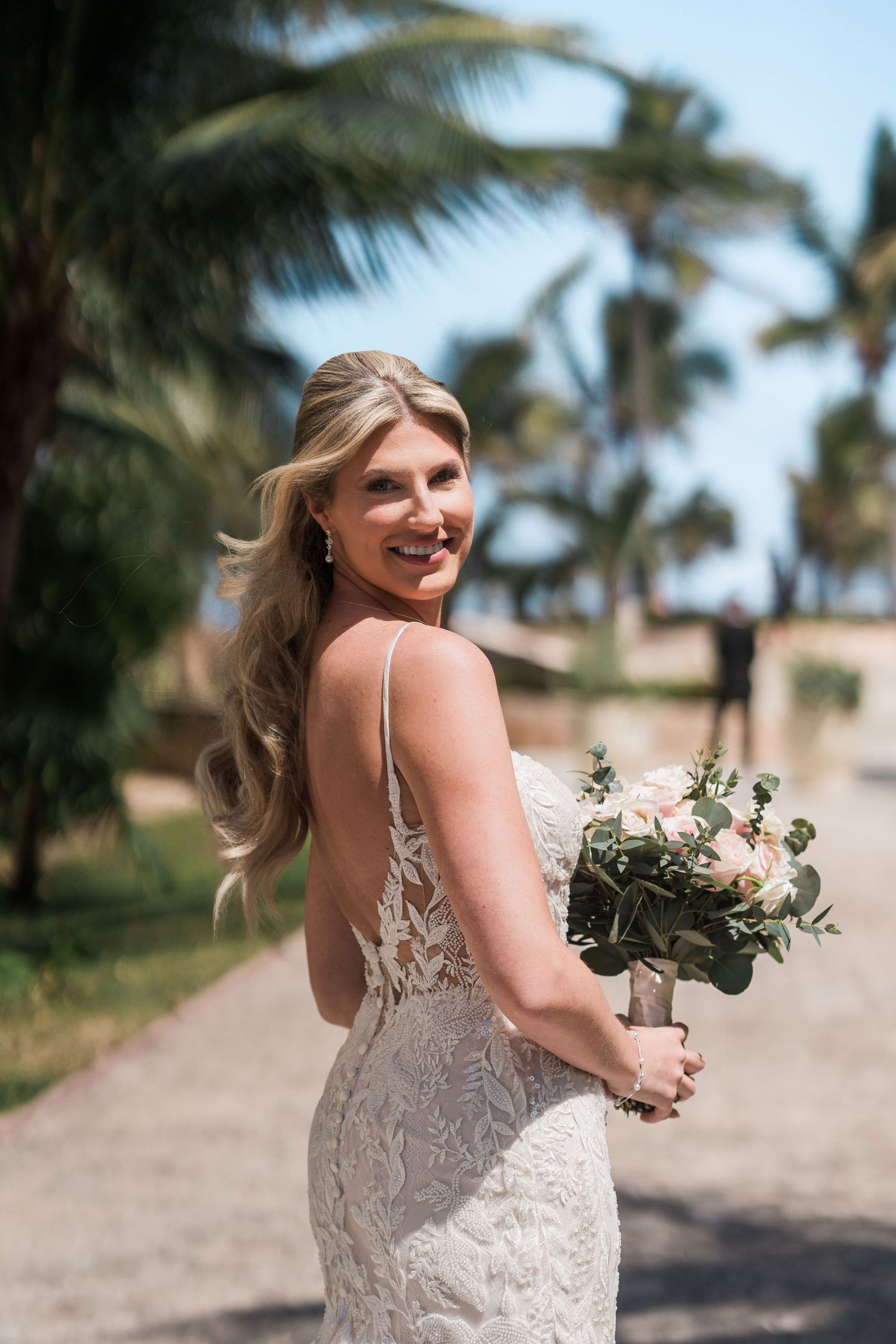 Smiling bride in lace backless gown holds bouquet of blush roses, posing outdoors near palm trees on her wedding day.