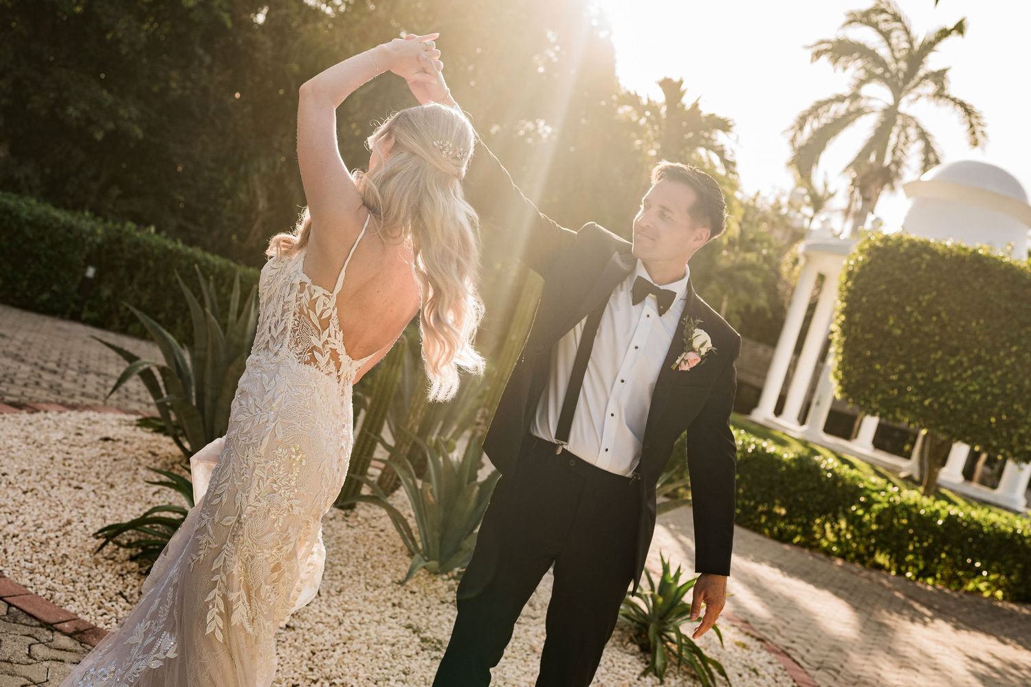 Bride and groom dancing outdoors in golden sunlight, woman in lace wedding gown, man in black tuxedo with boutonniere.