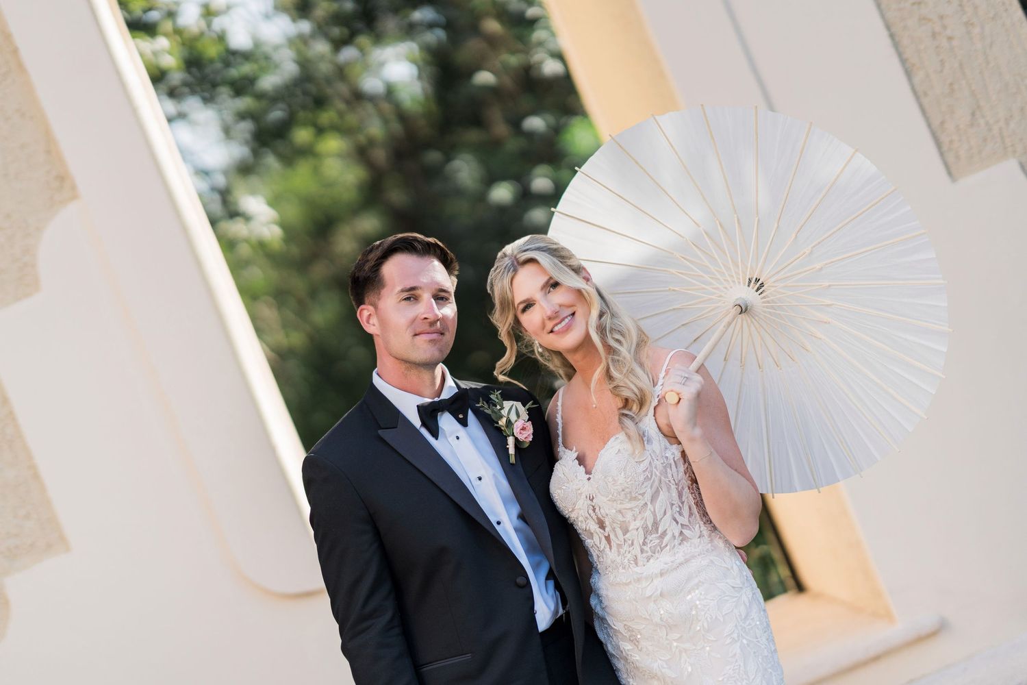 Bride and groom posing outdoors, bride holding white parasol, groom in black tuxedo, elegant wedding portrait.