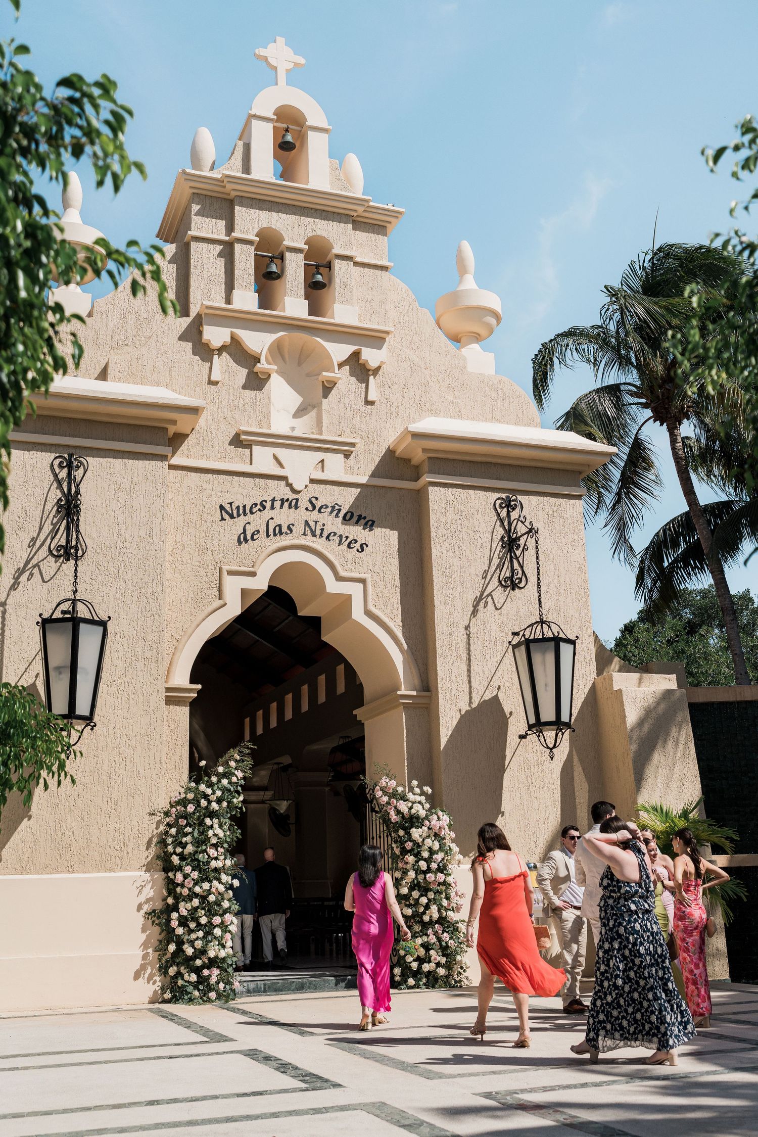 Guests arriving at Nuestra Señora de las Nieves chapel entrance adorned with floral arch under blue sky.