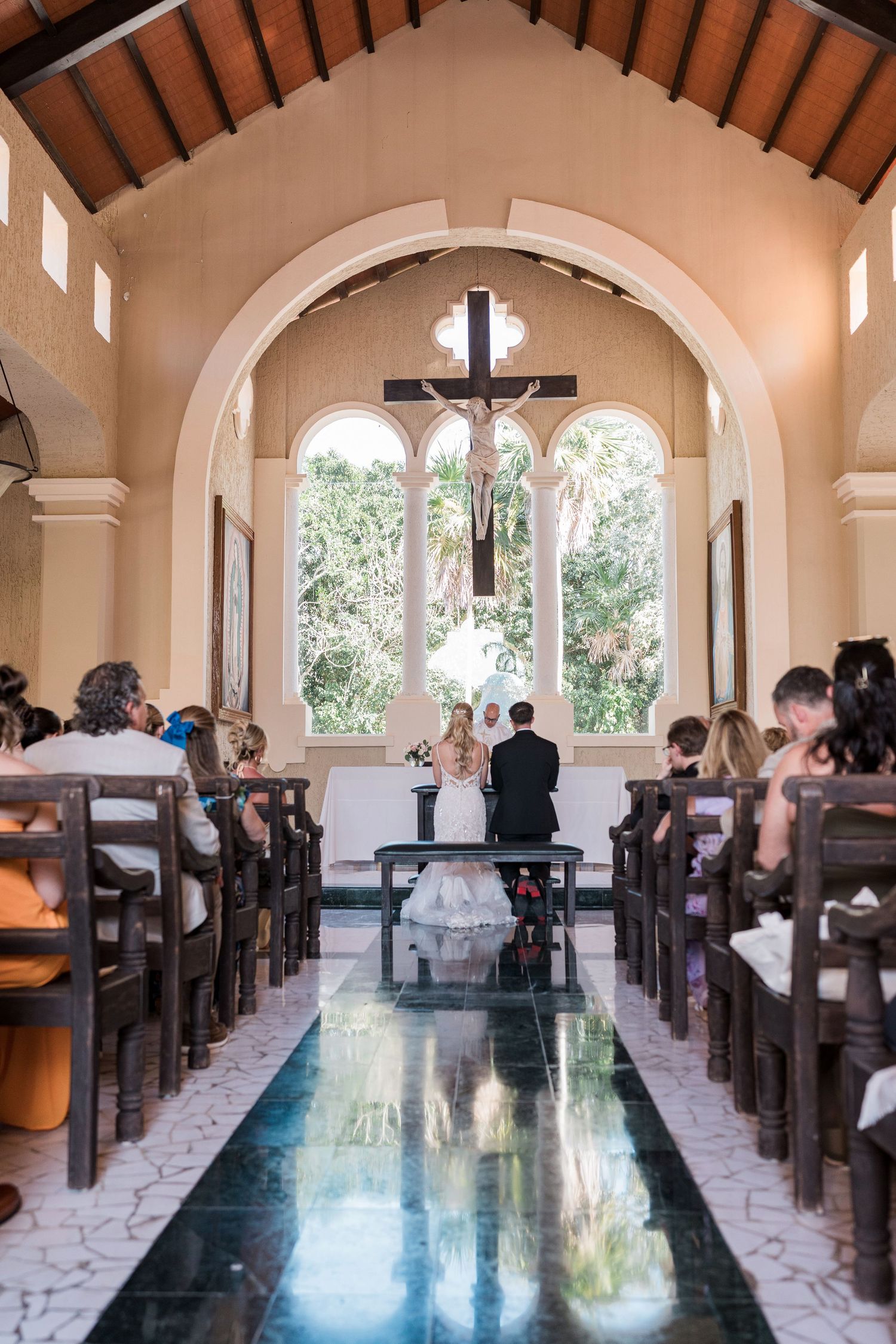 Elegant chapel wedding ceremony with couple at altar beneath a crucifix, guests seated in wooden pews, marble aisle.