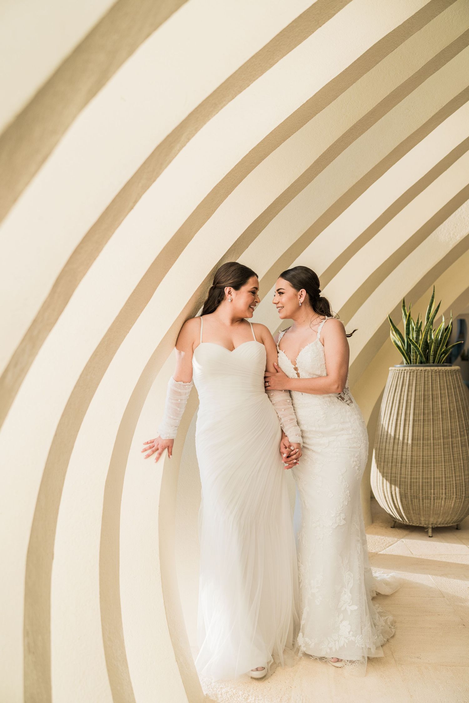 Two brides in white wedding gowns holding hands and smiling at each other against a curved striped wall.