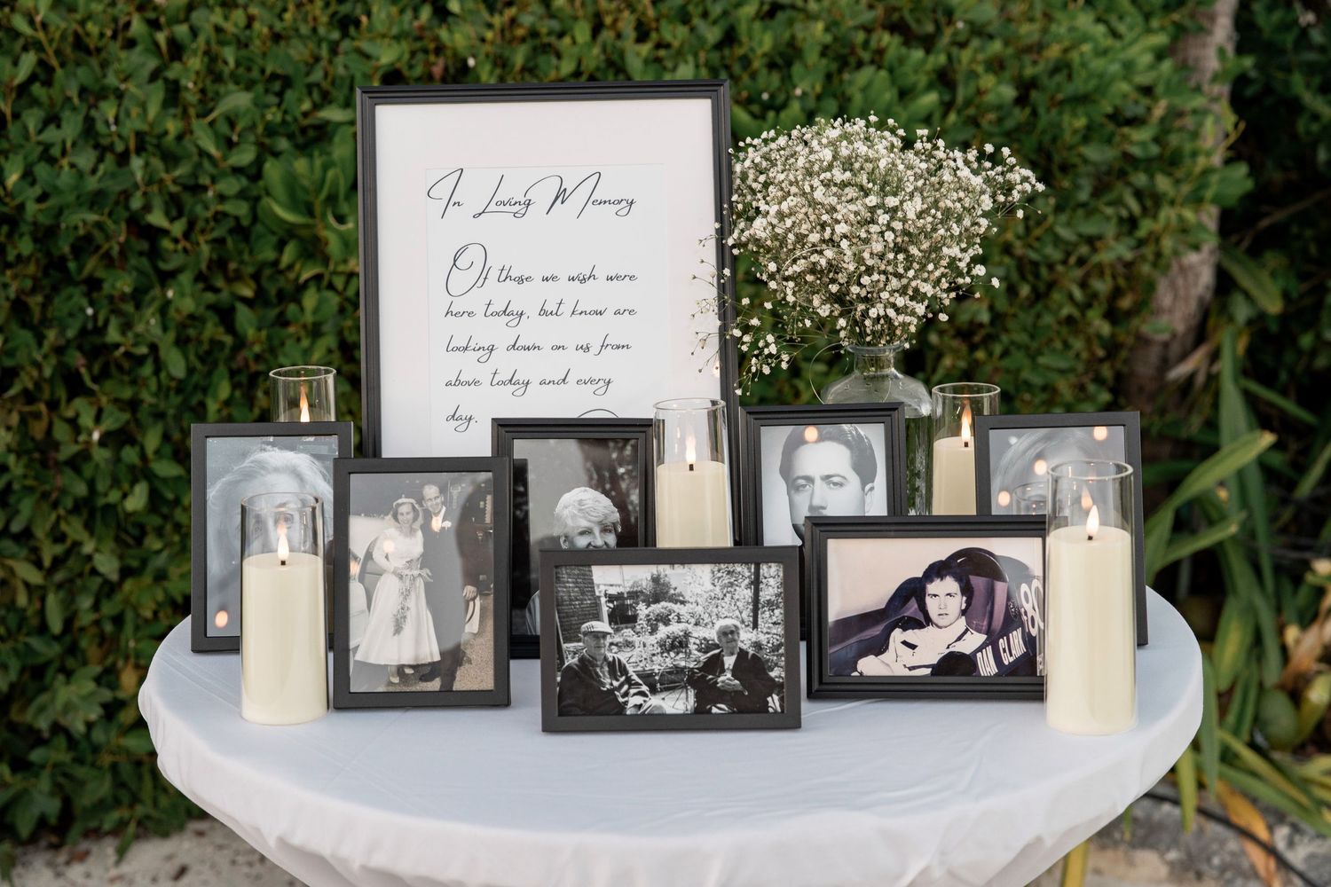 A memorial table display with framed black-and-white photos, white candles, and baby's breath flowers with an In Loving Memory sign.