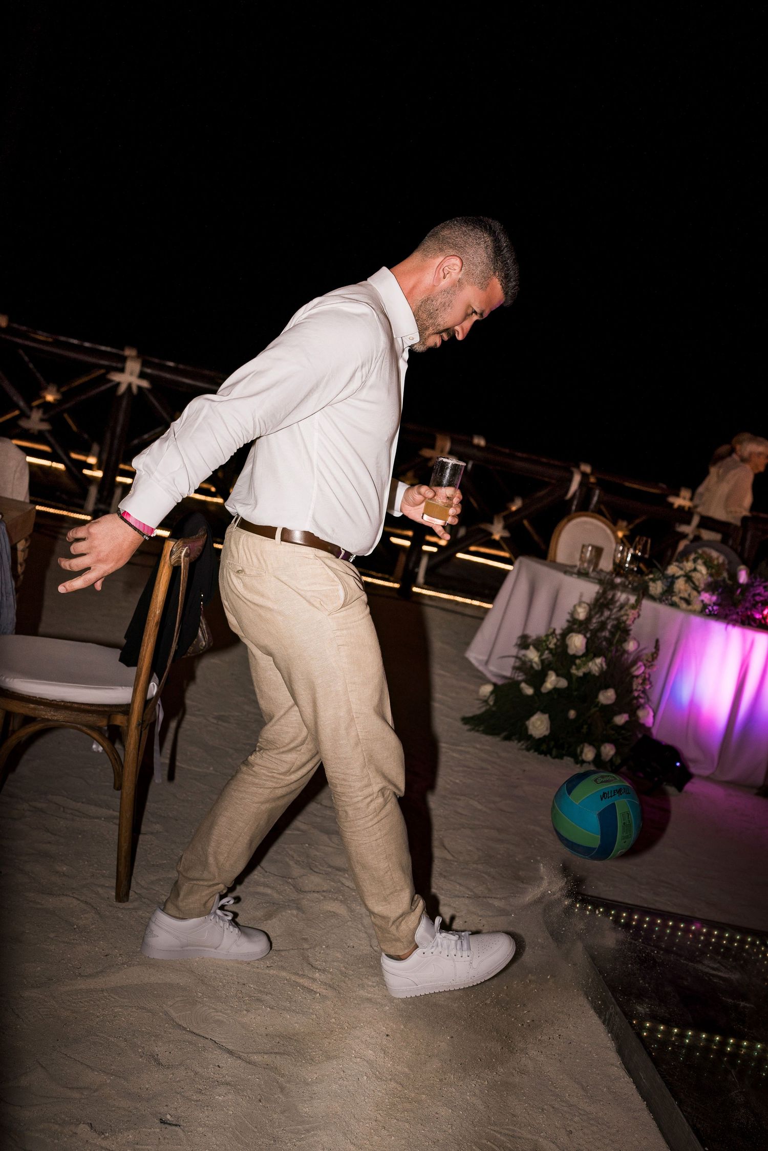 Man in white shirt and khaki pants dancing on sand at an outdoor evening event holding a drink.
