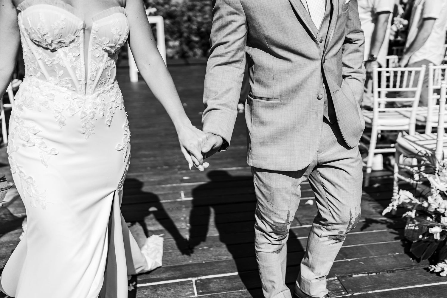 Black and white photo of bride and groom holding hands walking down the aisle at outdoor wedding ceremony.
