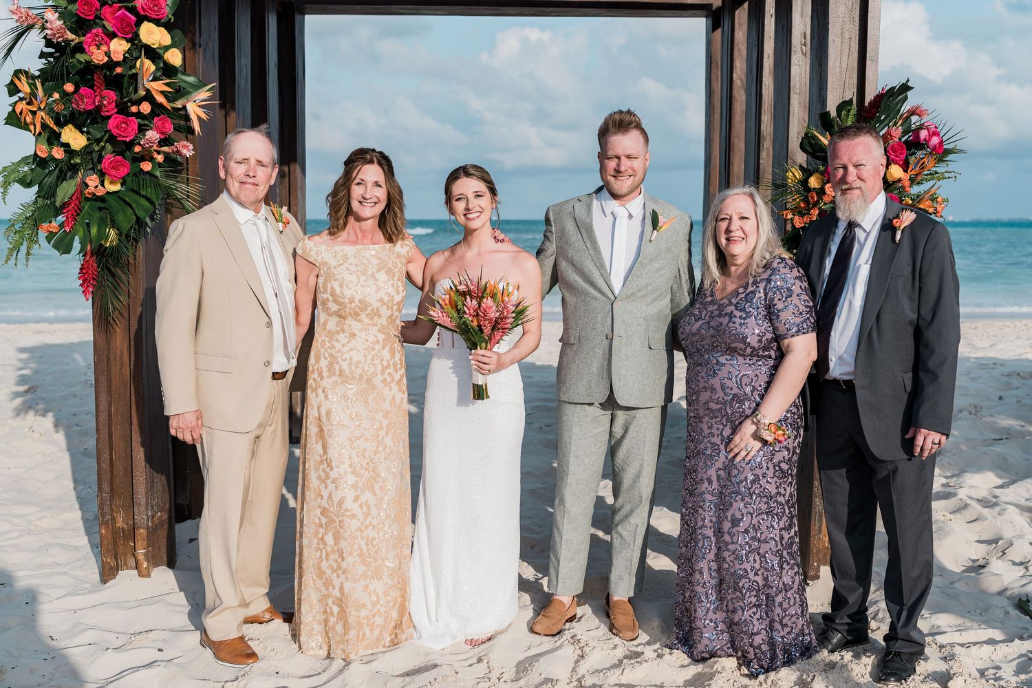 Wedding party posing on a beach ceremony with tropical floral arrangements, bride holding bouquet in elegant outdoor setting.