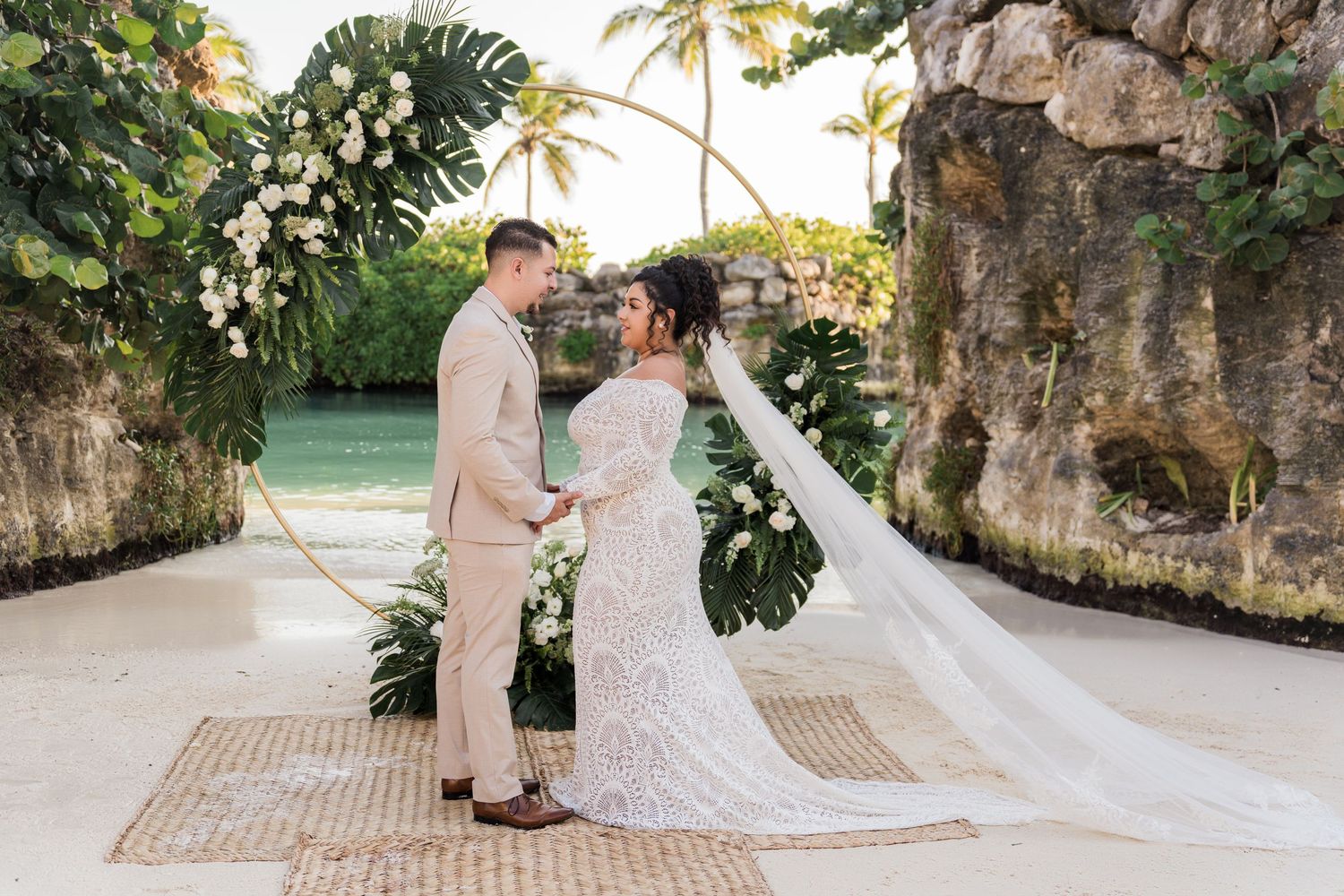 Xcaret Arte Wedding - Bride and groom hold hands at tropical beach wedding altar with floral arch, palm trees, and turquoise water backdrop.
