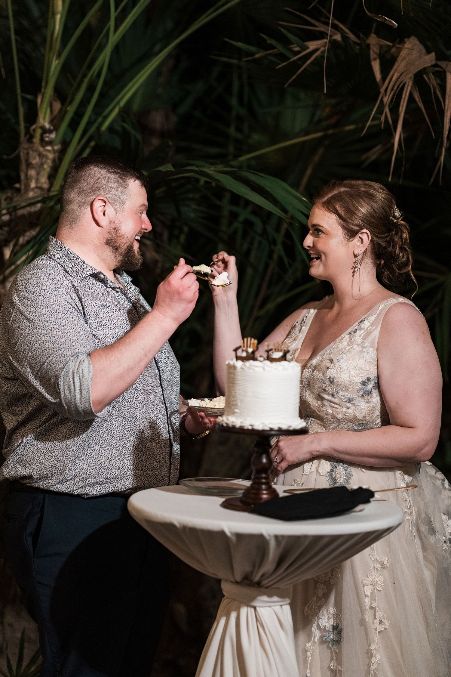 Secrets Akumal Wedding - Bride and groom laughing while feeding each other wedding cake at tropical outdoor reception.
