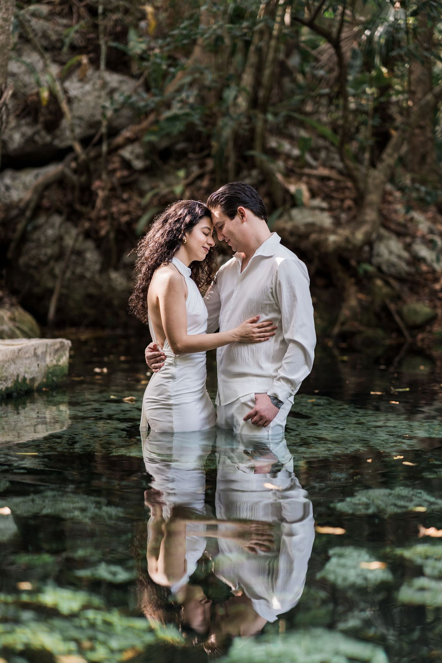 Couple embracing in a serene forest cenote, wearing white outfits, with their reflection visible in the clear water.