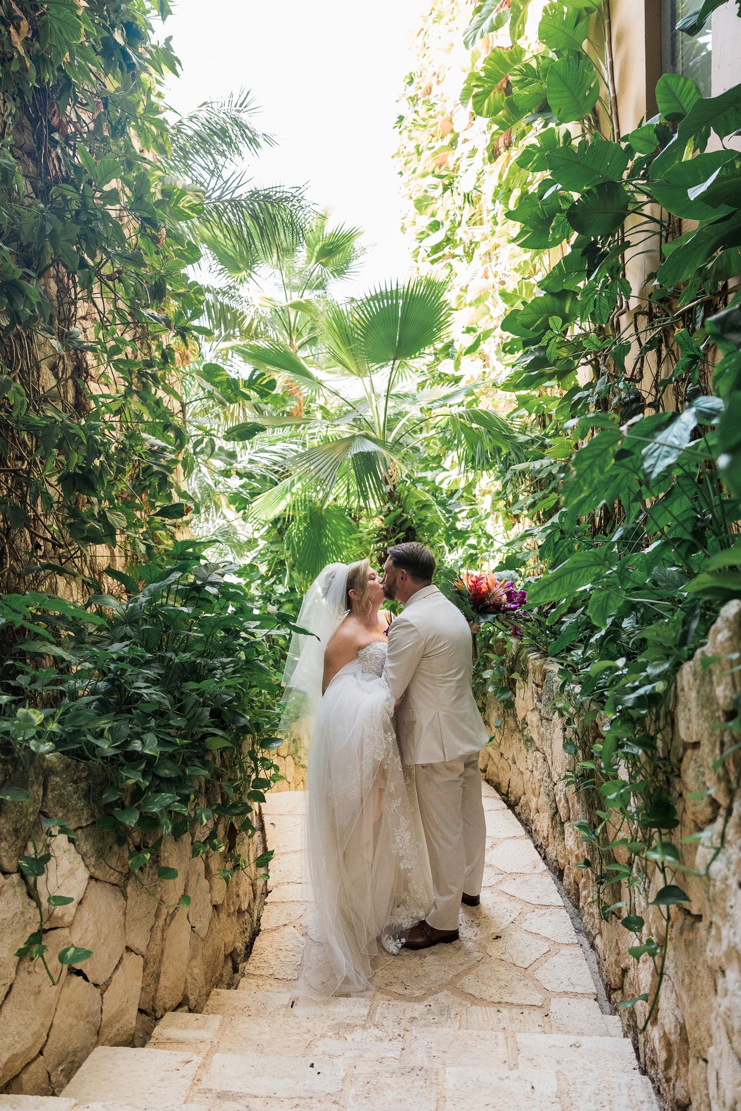 Bride and groom share a romantic kiss on stone steps surrounded by lush tropical greenery at an outdoor wedding.