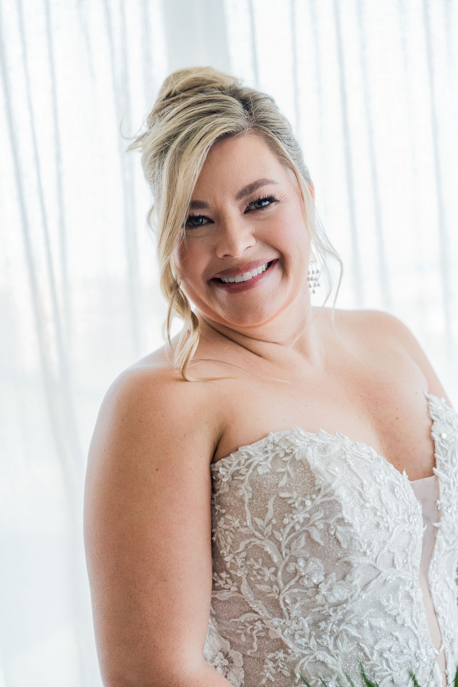 Smiling bride in strapless lace wedding gown with updo hairstyle and chandelier earrings posing near bright window.