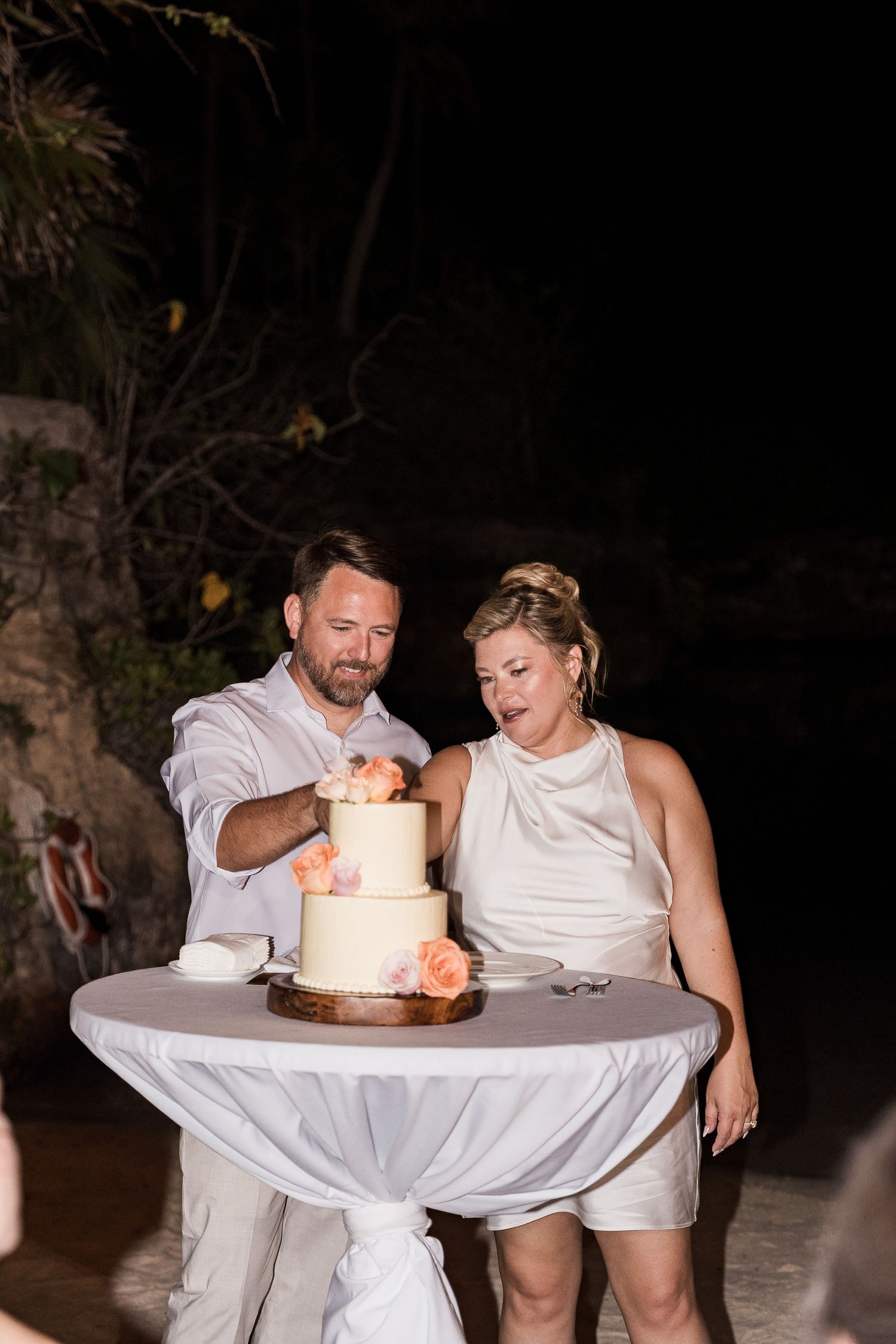 Couple cutting a two-tier white wedding cake with peach floral decorations at an outdoor evening reception.