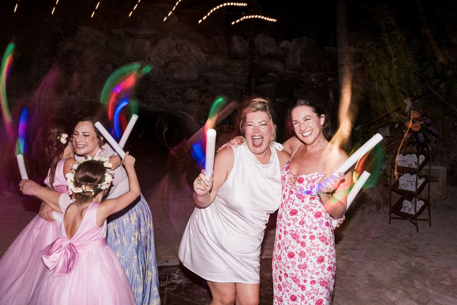 Women in formal wear dance joyfully at an outdoor wedding reception holding colorful glow sticks at night.