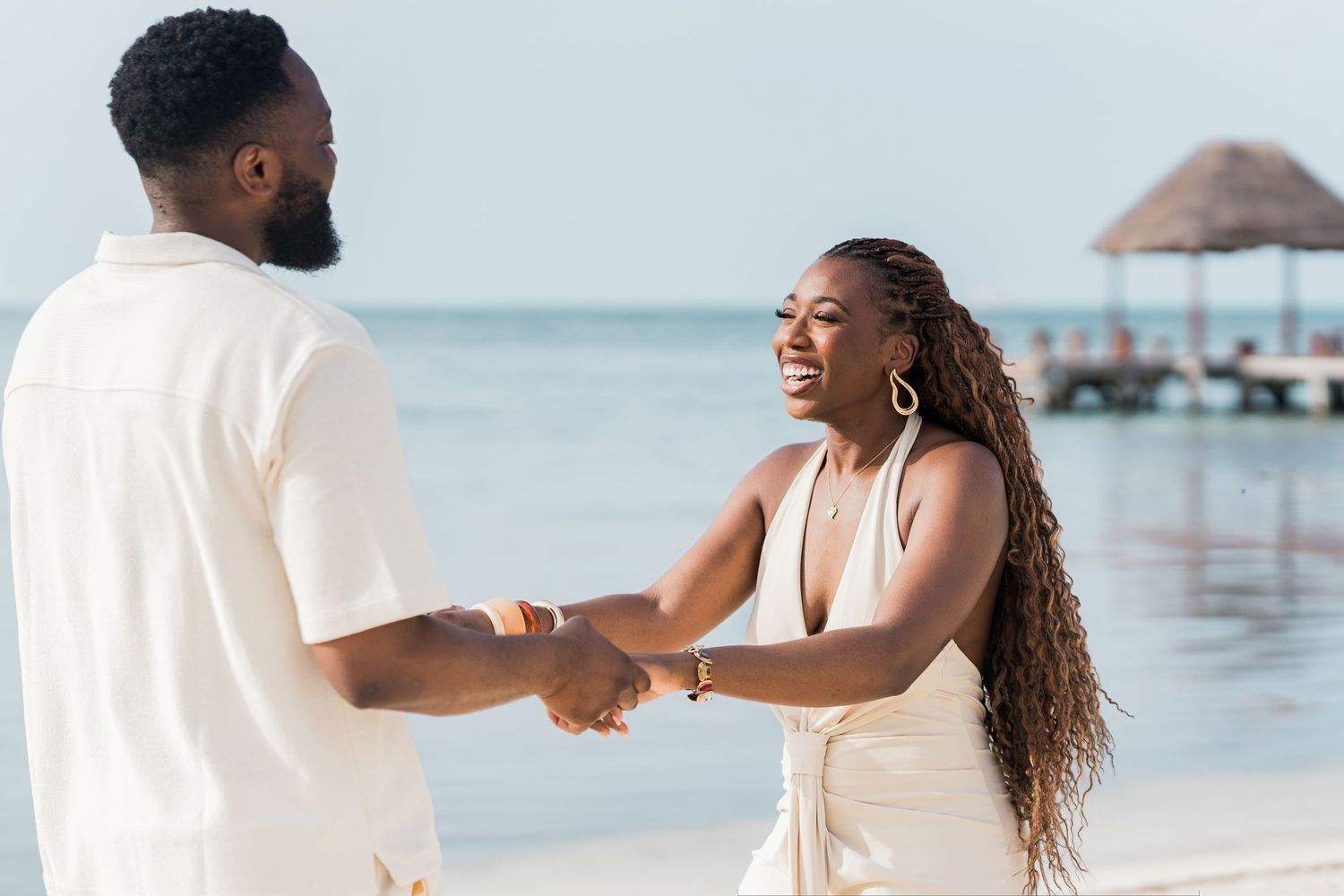 Couple holding hands and smiling on a tropical beach, dressed in white outfits with ocean in background.