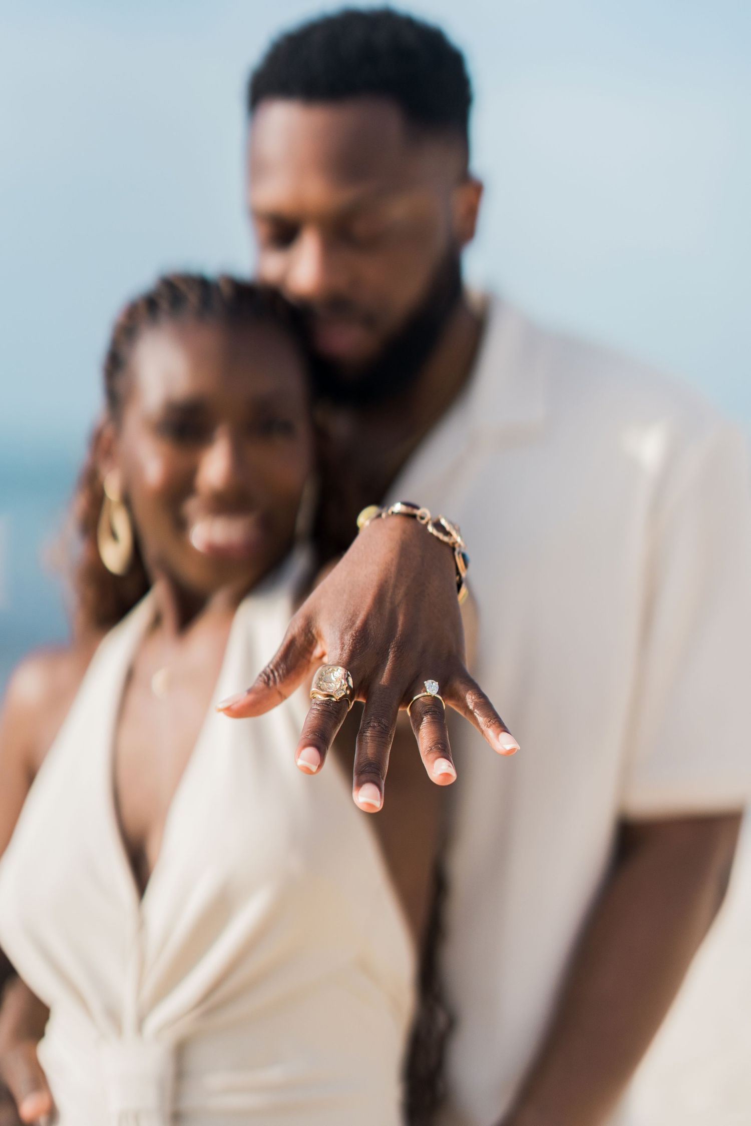 An engaged Black couple displays a sparkling diamond engagement ring on a bright beach, both dressed in white outfits.