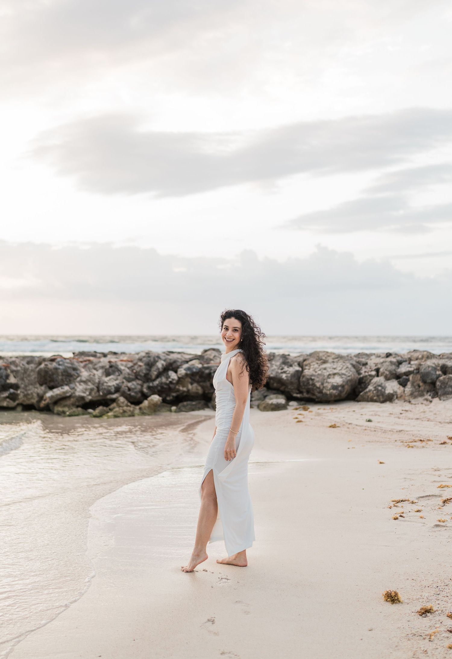 Woman in white dress walking barefoot on sandy beach with rocky shoreline and cloudy sky backdrop.