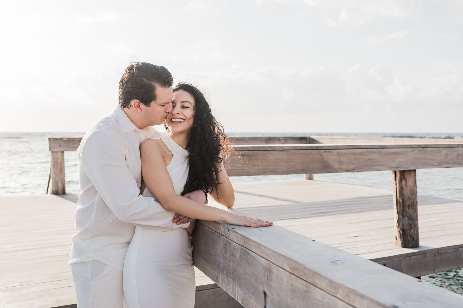 Couple embracing on a wooden pier by the ocean during golden hour, wearing white wedding attire.