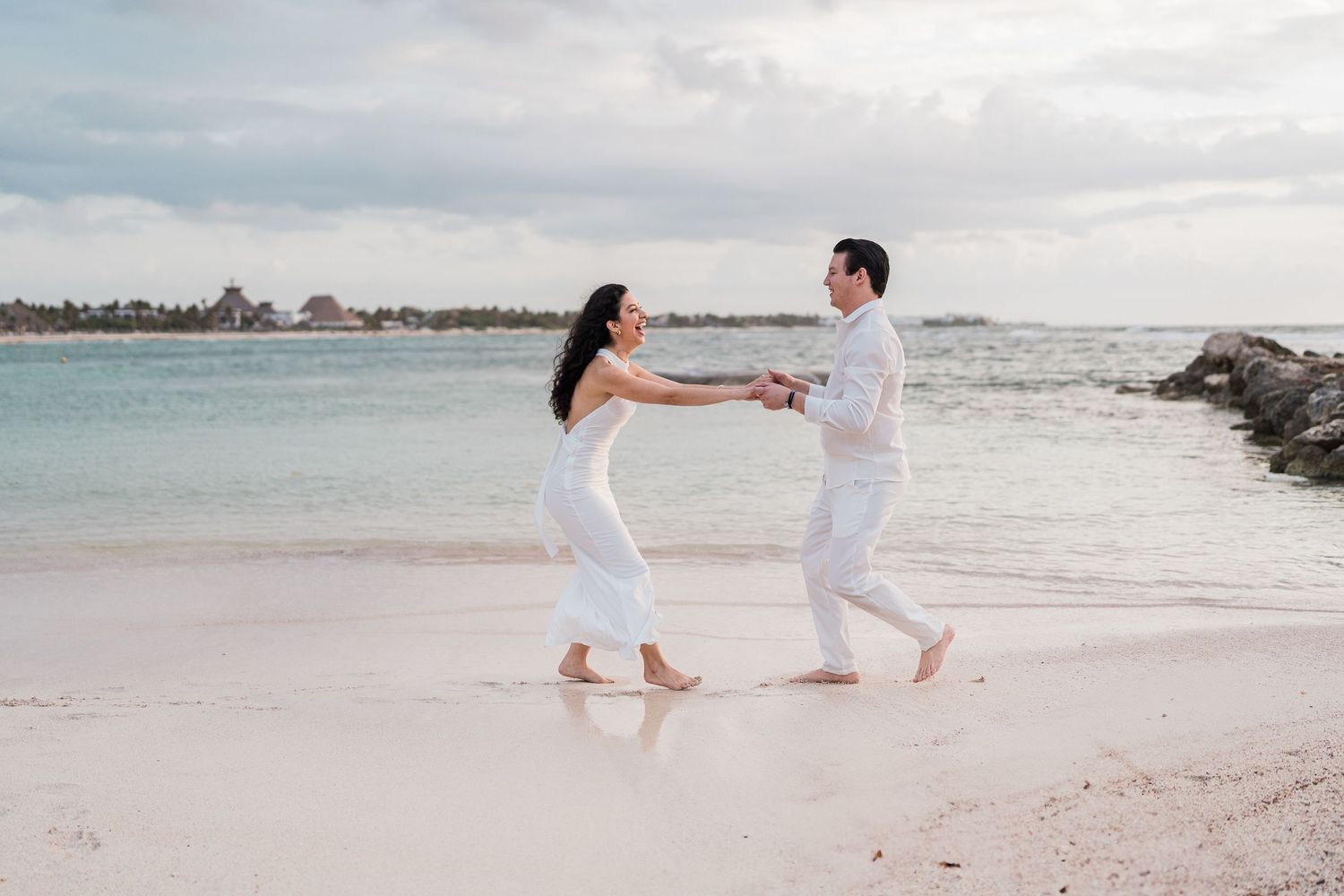 Couple in white outfits holding hands and dancing barefoot on a tropical beach with calm turquoise water.