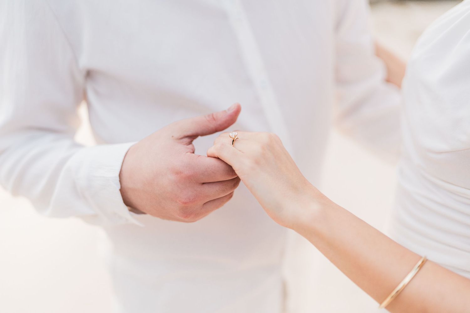 Couple exchanging wedding rings outdoors, groom in white shirt placing gold ring on bride's finger.