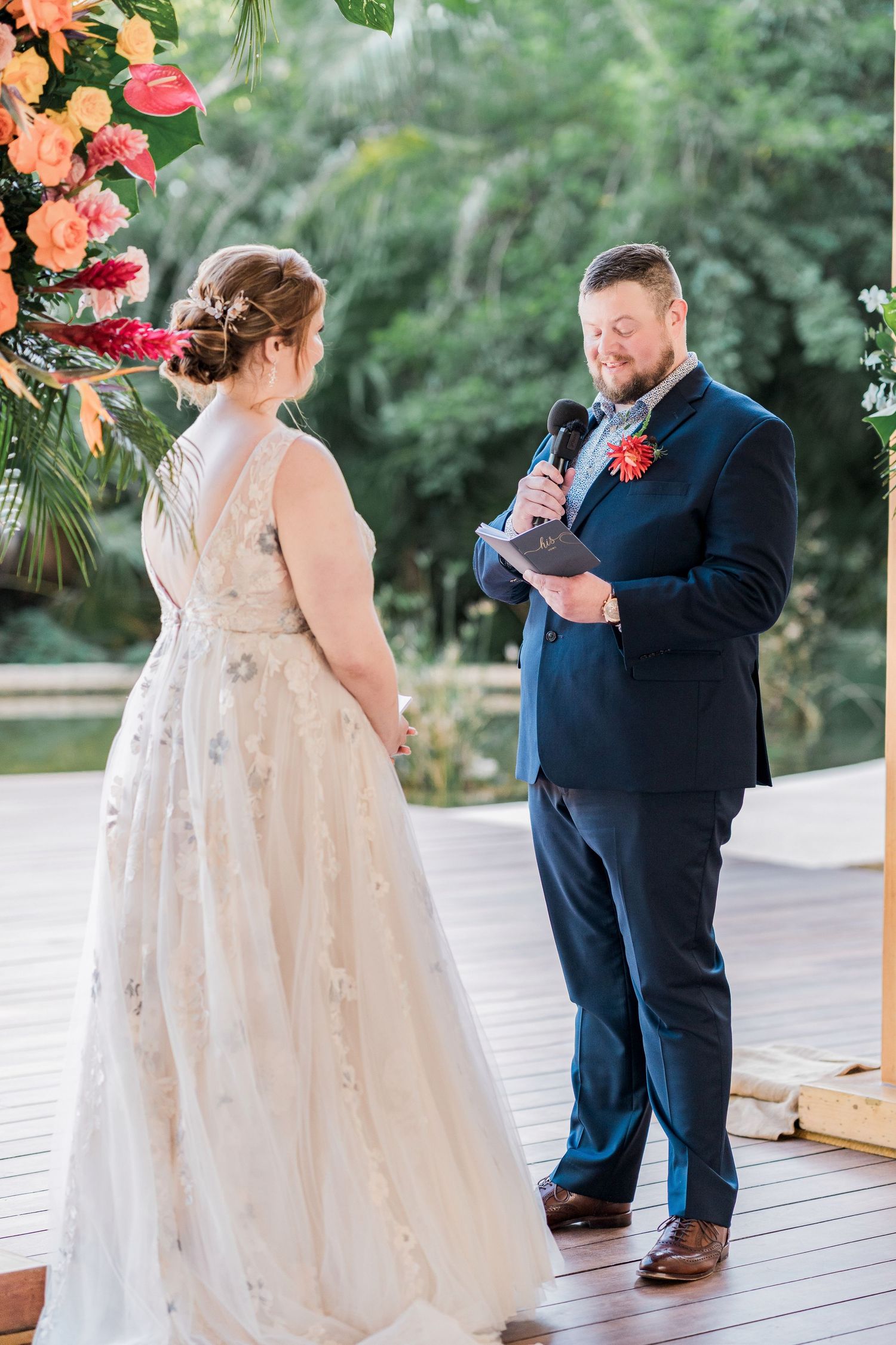 Secrets Akumal Wedding - Groom reads wedding vows to bride in floral wedding dress on tropical outdoor deck during ceremony.