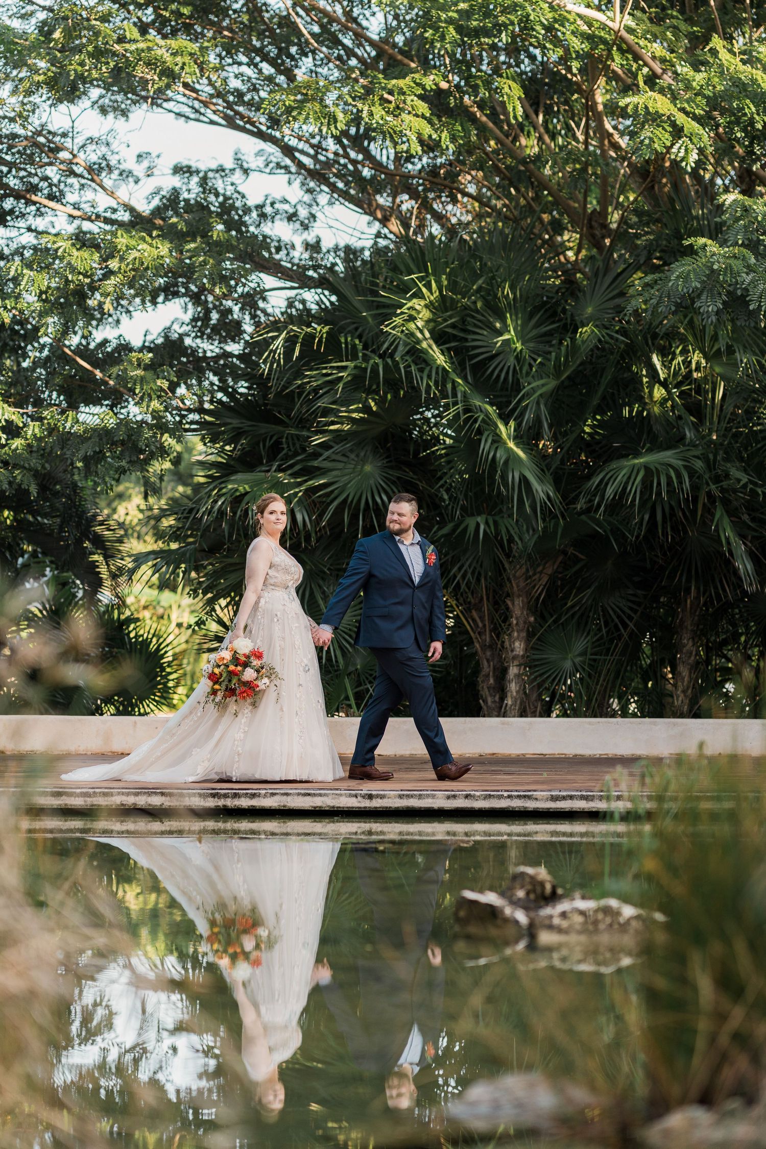 Secrets Akumal Wedding Bride and groom walk hand-in-hand beside a reflective pond surrounded by lush tropical greenery on their wedding day.