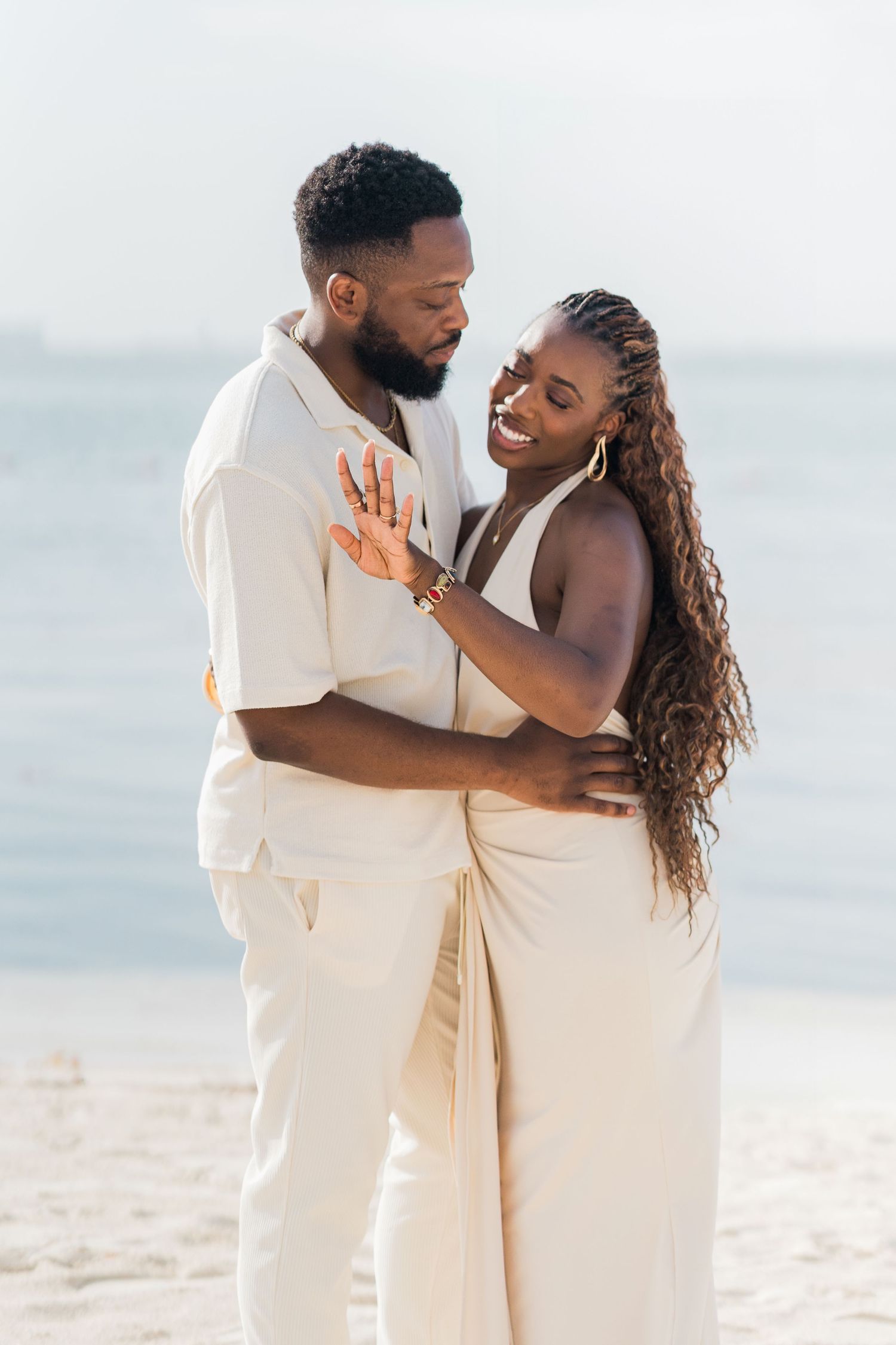 Happy couple embraces on a sunny beach, both wearing matching cream outfits, smiling and holding each other close.