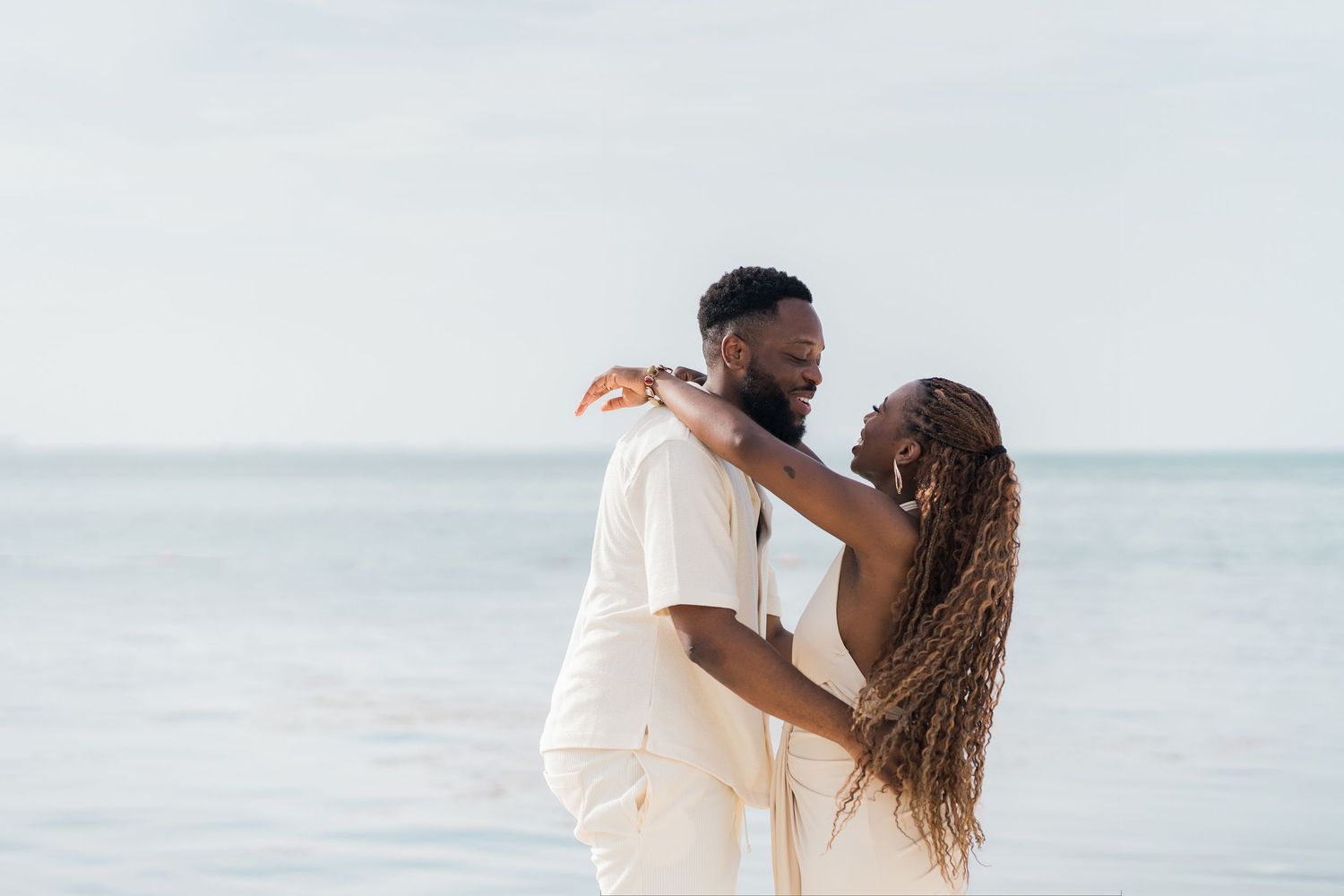 Couple embracing romantically on a beach, wearing white outfits, with calm ocean waves in the background.
