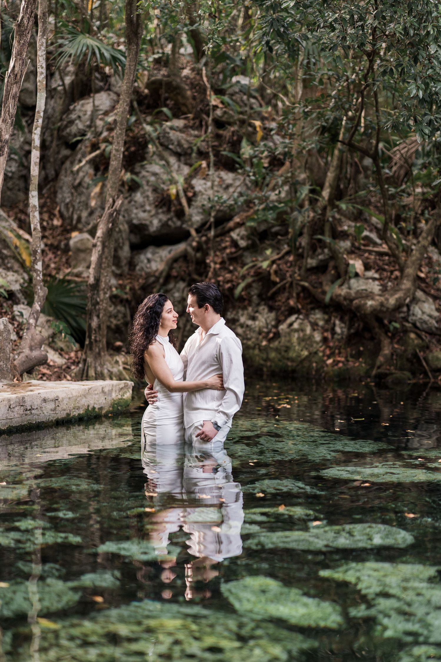Couple embracing in a lush jungle cenote, standing in clear green water surrounded by rocks and tropical trees.