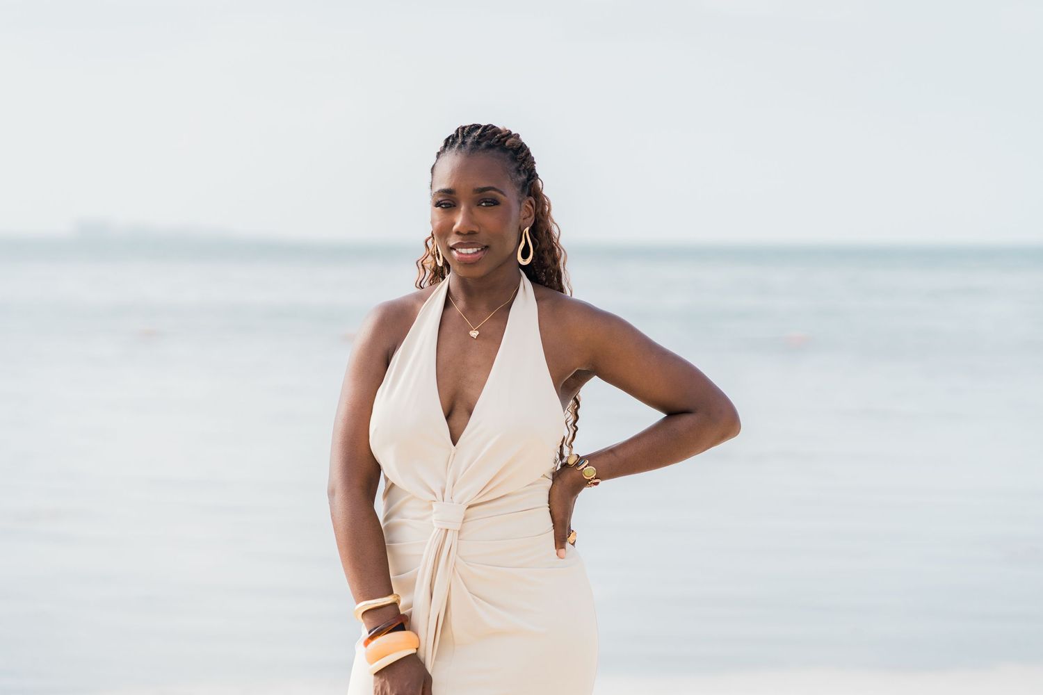 Smiling woman in cream halter dress posing on a beach with ocean in the background.