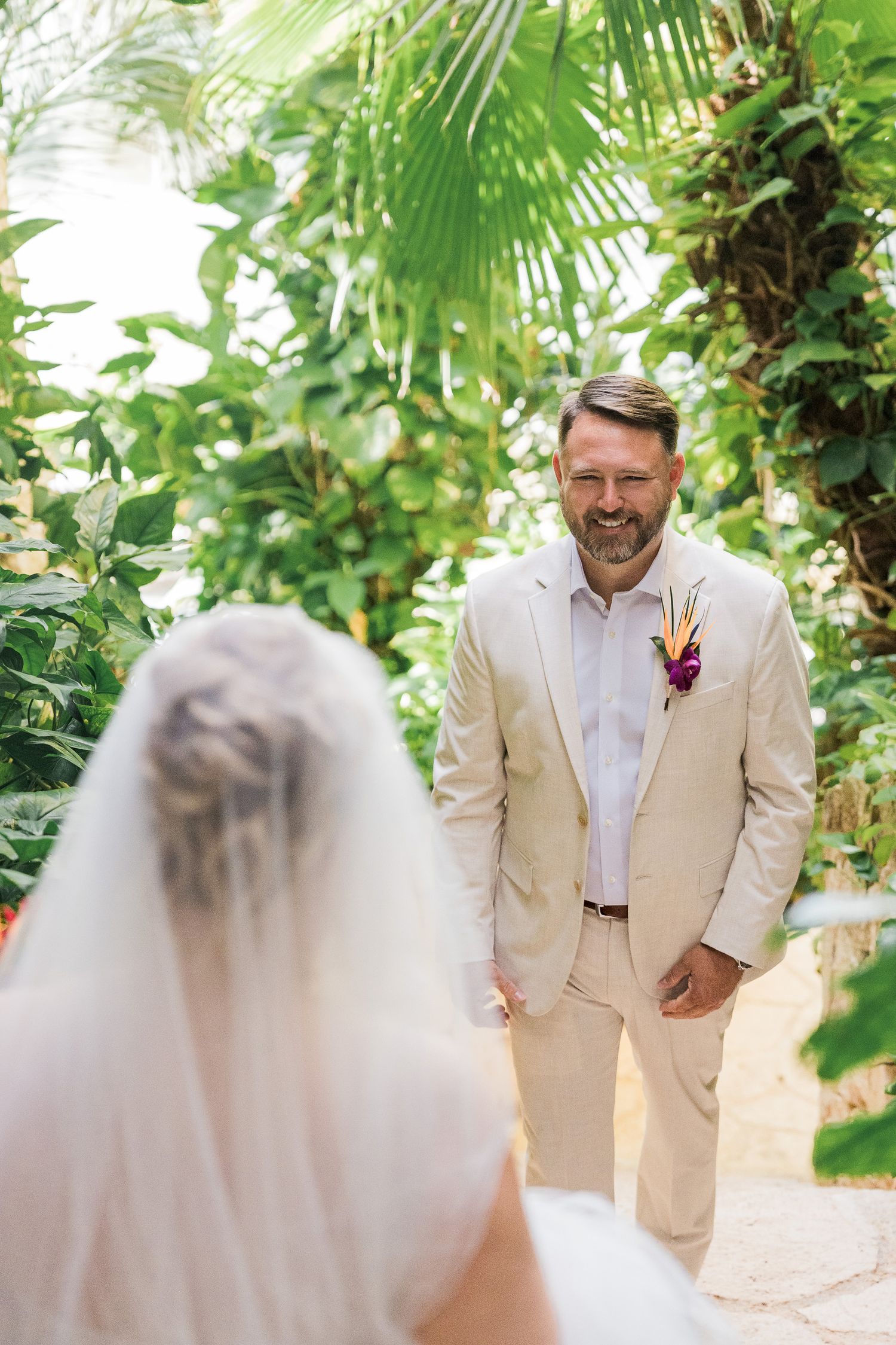 Hotel Xcaret México Wedding -Groom in beige suit smiling at bride during outdoor tropical wedding ceremony surrounded by lush green foliage.