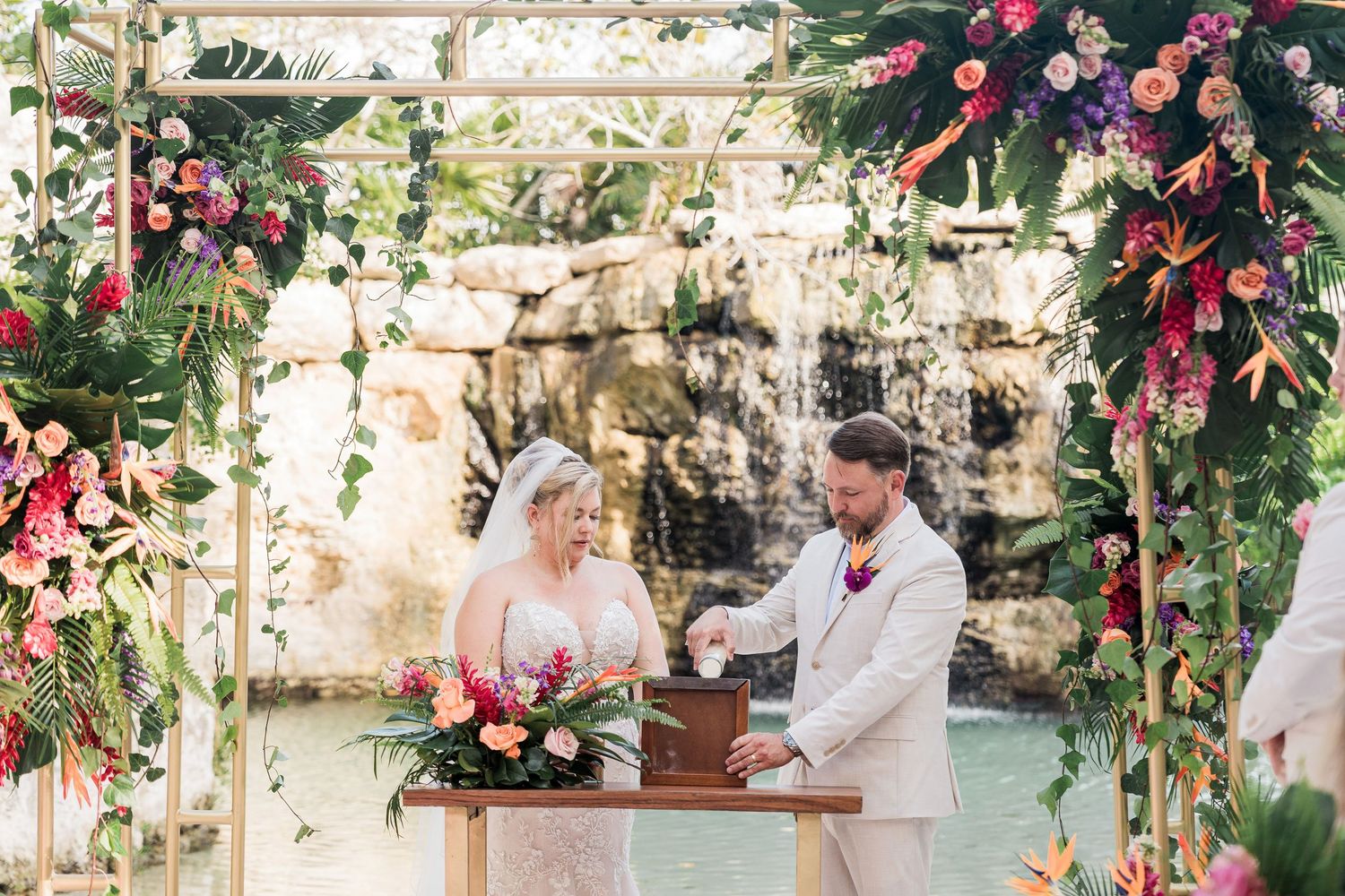 Hotel Xcaret México Wedding  - Bride and groom signing wedding documents under floral arch with waterfall backdrop and tropical flowers.