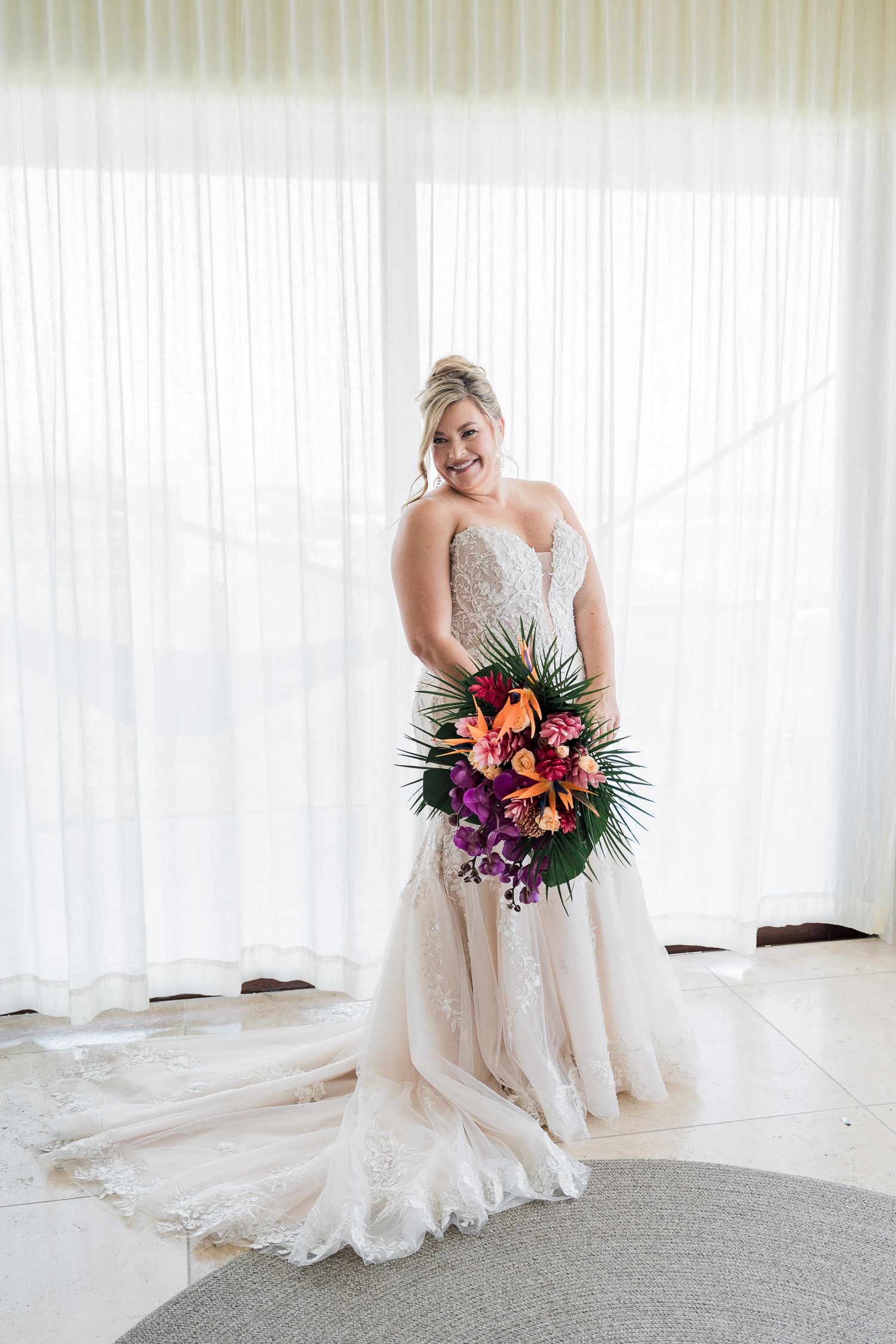 Bride in a beaded strapless wedding gown holding a vibrant tropical bouquet with purple and orange flowers, standing before sheer curtains.