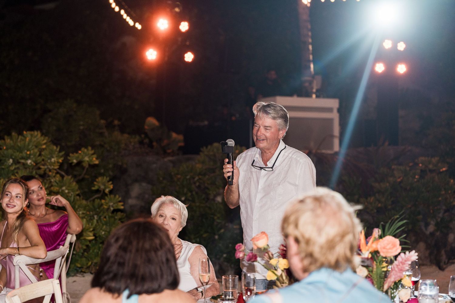 Man giving a speech with a microphone at an outdoor evening wedding reception with guests seated at decorated tables.