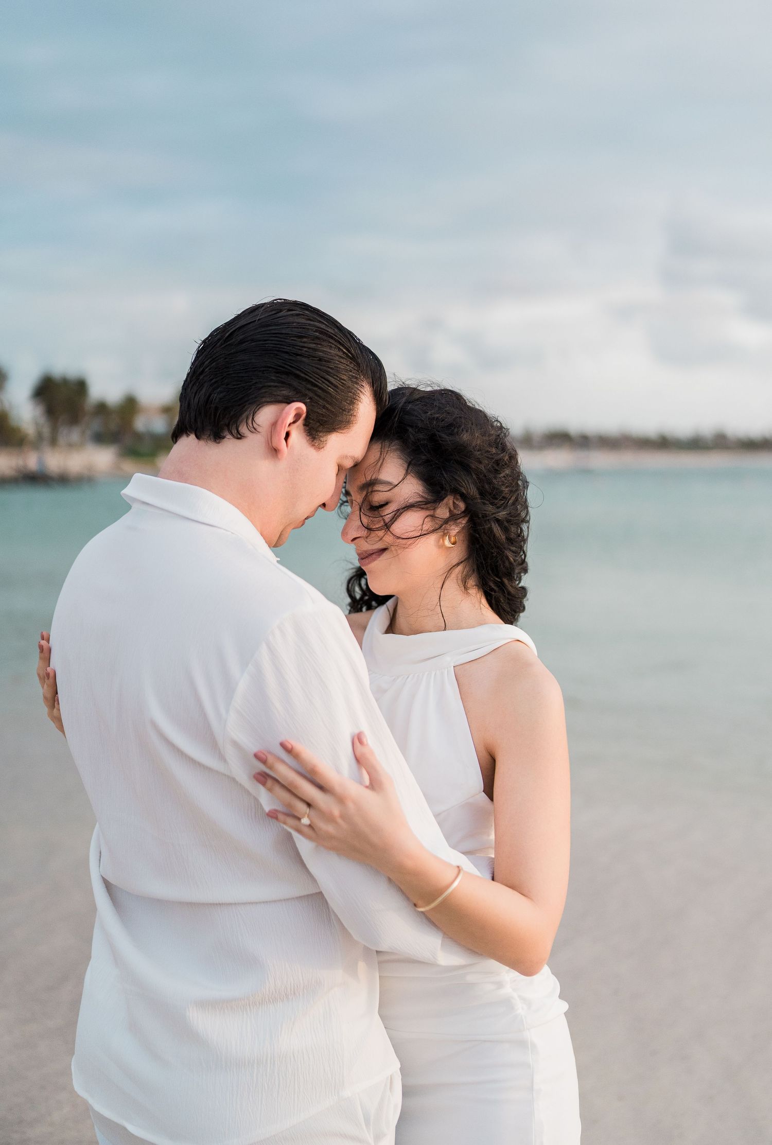 Couple in white outfits embracing on a tropical beach, sharing a romantic moment by the turquoise water.
