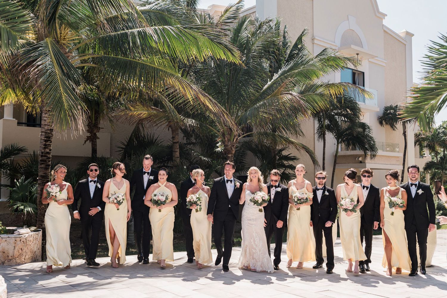 Wedding party in formal attire poses outdoors with palm trees, bridesmaids in gold gowns and groomsmen in black tuxedos.