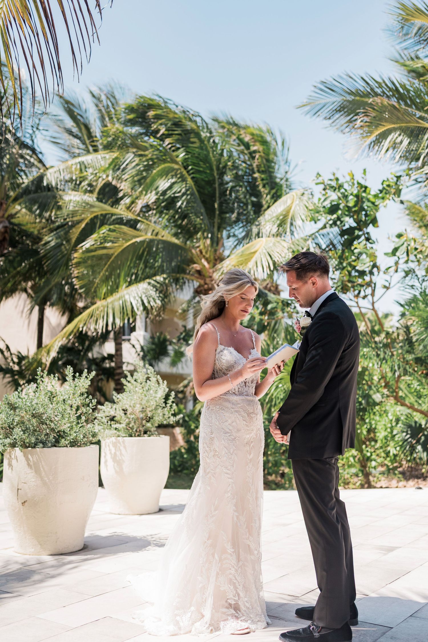 Bride and groom exchange rings during outdoor tropical wedding ceremony with palm trees and blue sky.