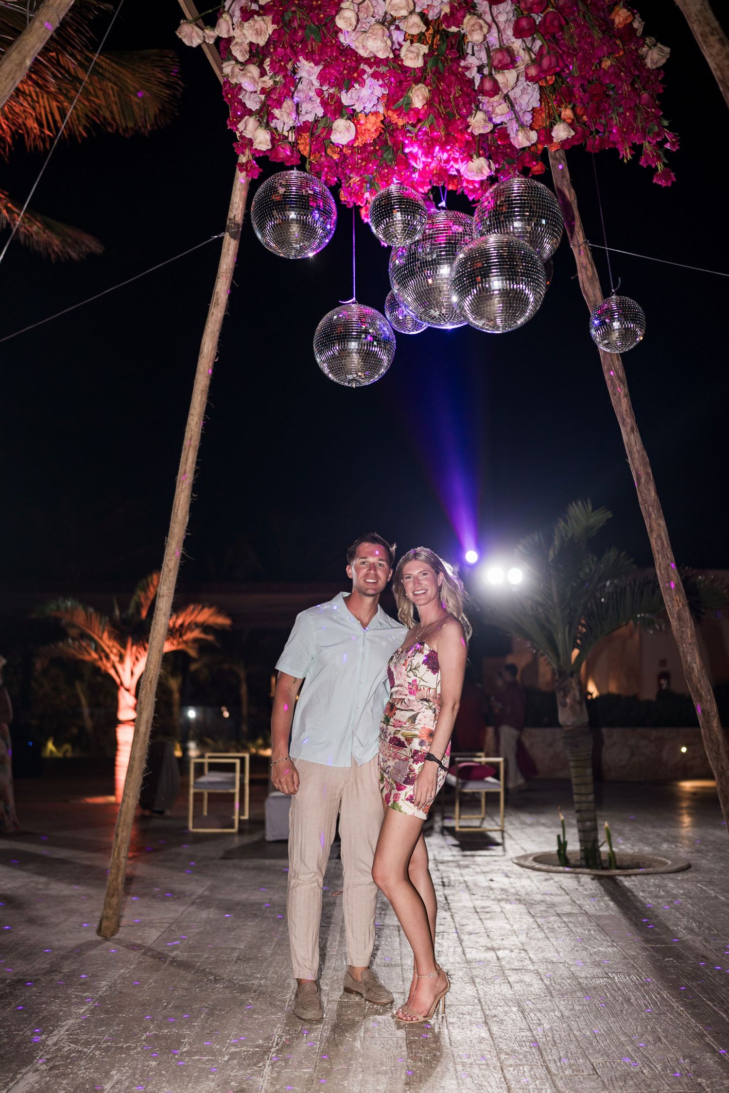 Couple posing under floral arch with disco balls at night outdoor event venue.