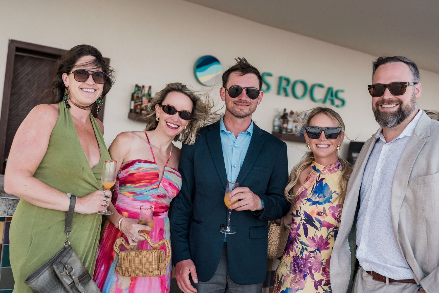 Group of five adults smiling and holding drinks outside Las Rocas resort on a sunny day.