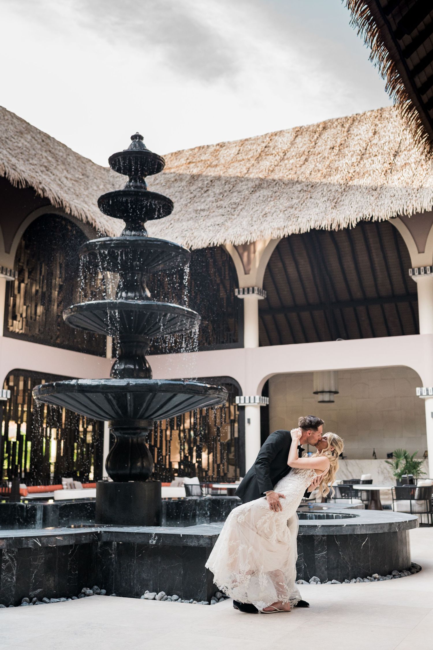 Bride and groom share a romantic dip kiss beside a black tiered fountain at an elegant resort courtyard.