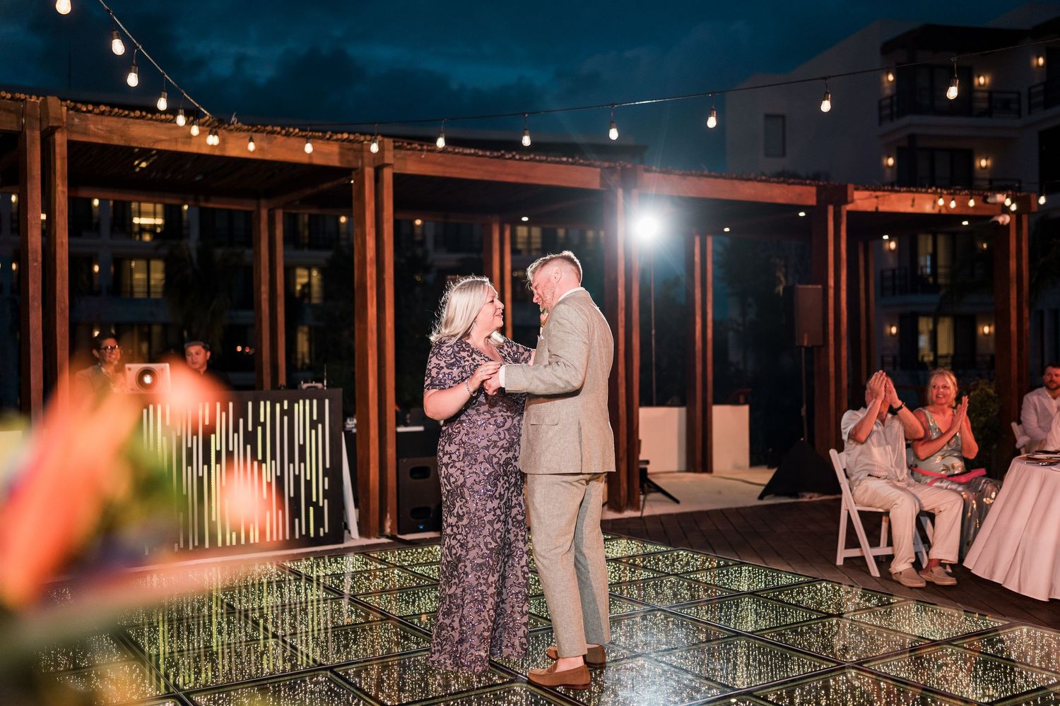 Couple shares first dance on reflective outdoor floor at elegant evening wedding reception with string lights.