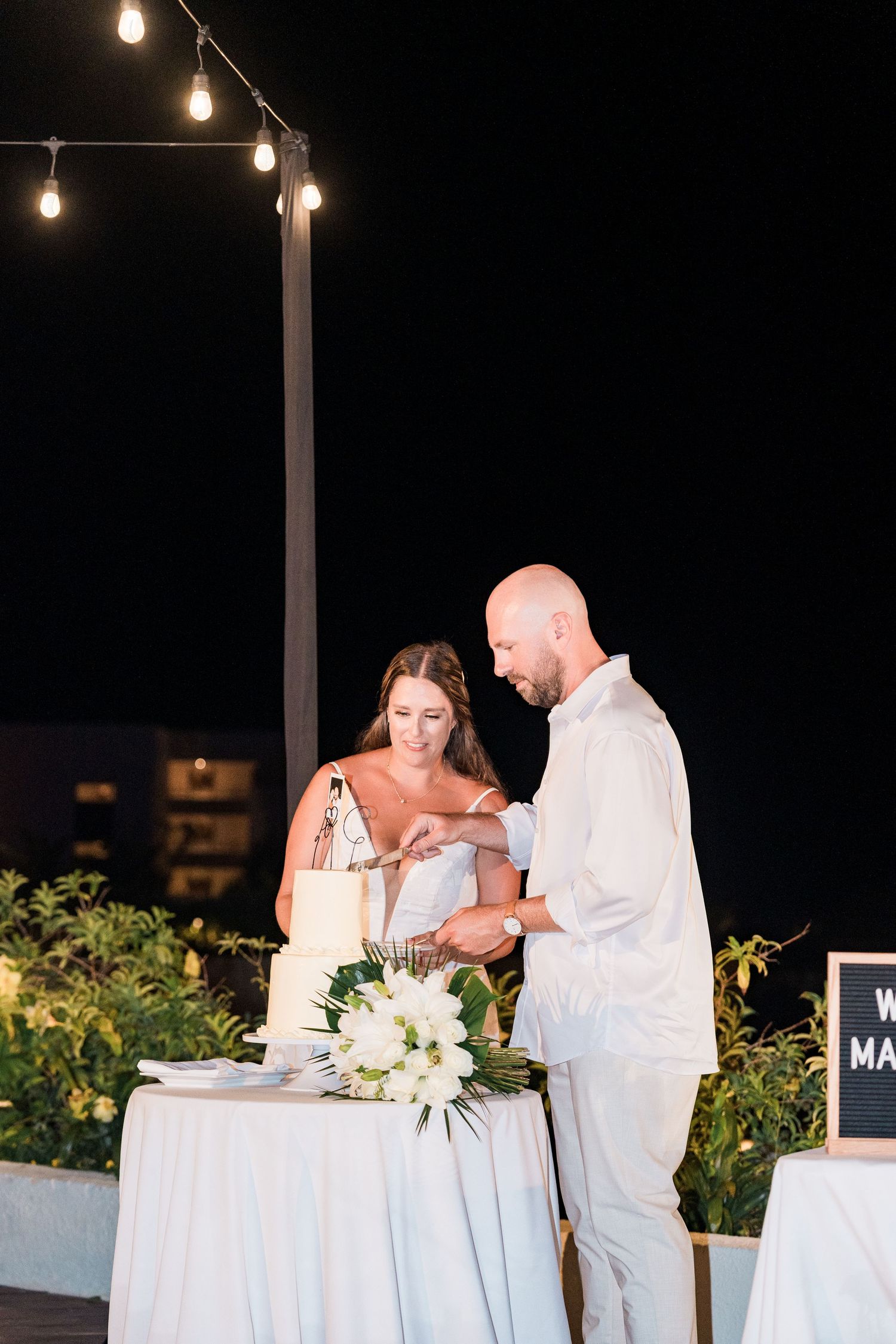 Couple cutting white wedding cake outdoors at night under string lights at elegant reception.