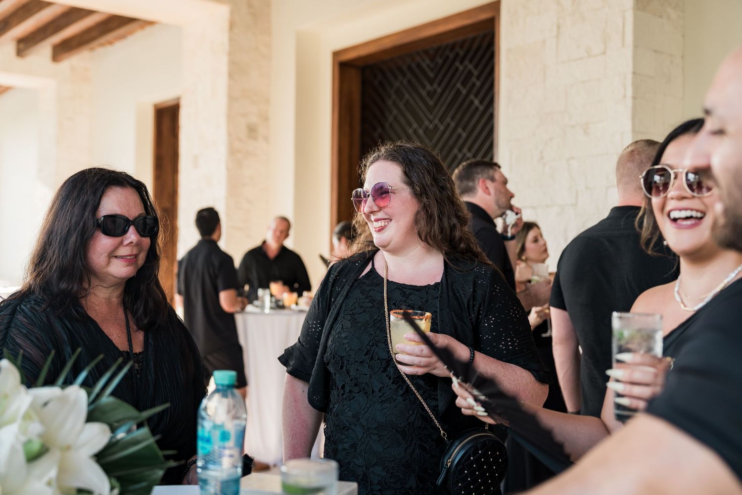 Guests in black attire laugh and enjoy drinks at an elegant outdoor wedding reception.