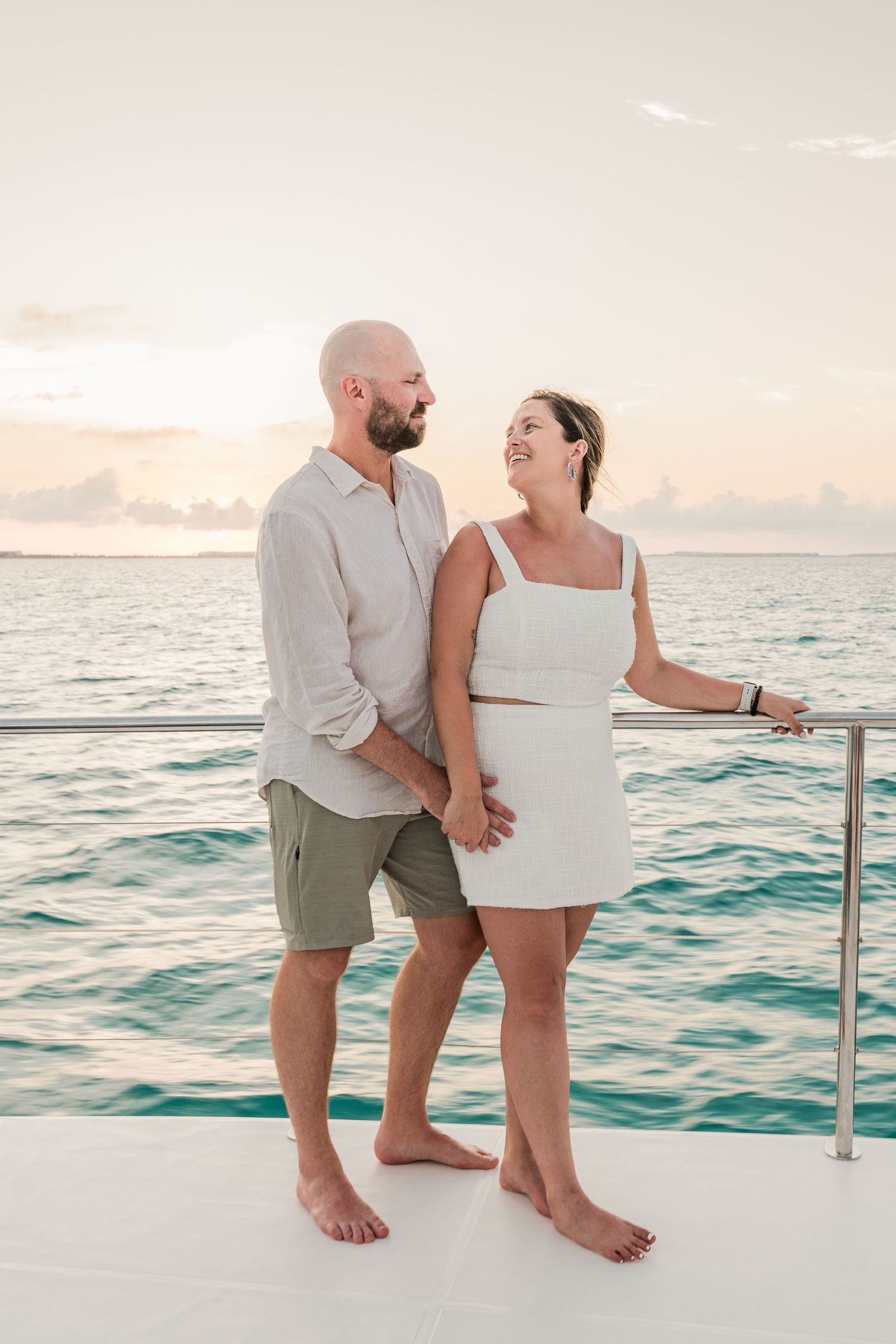 Couple holding hands on a yacht deck at sunset, wearing white and green outfits with turquoise ocean behind them.